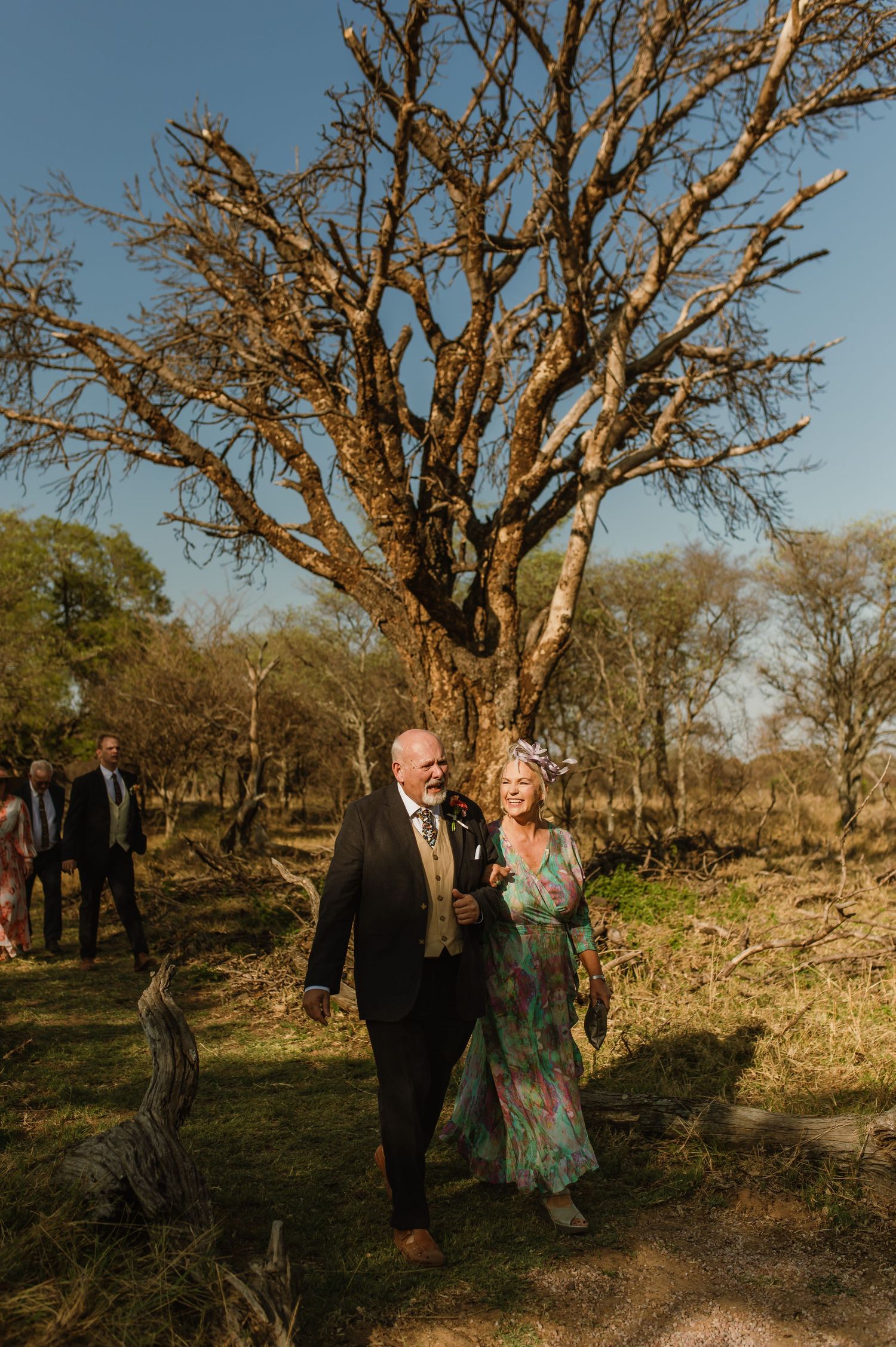 Couple walks through rugged terrain with gnarled trees during sunset.