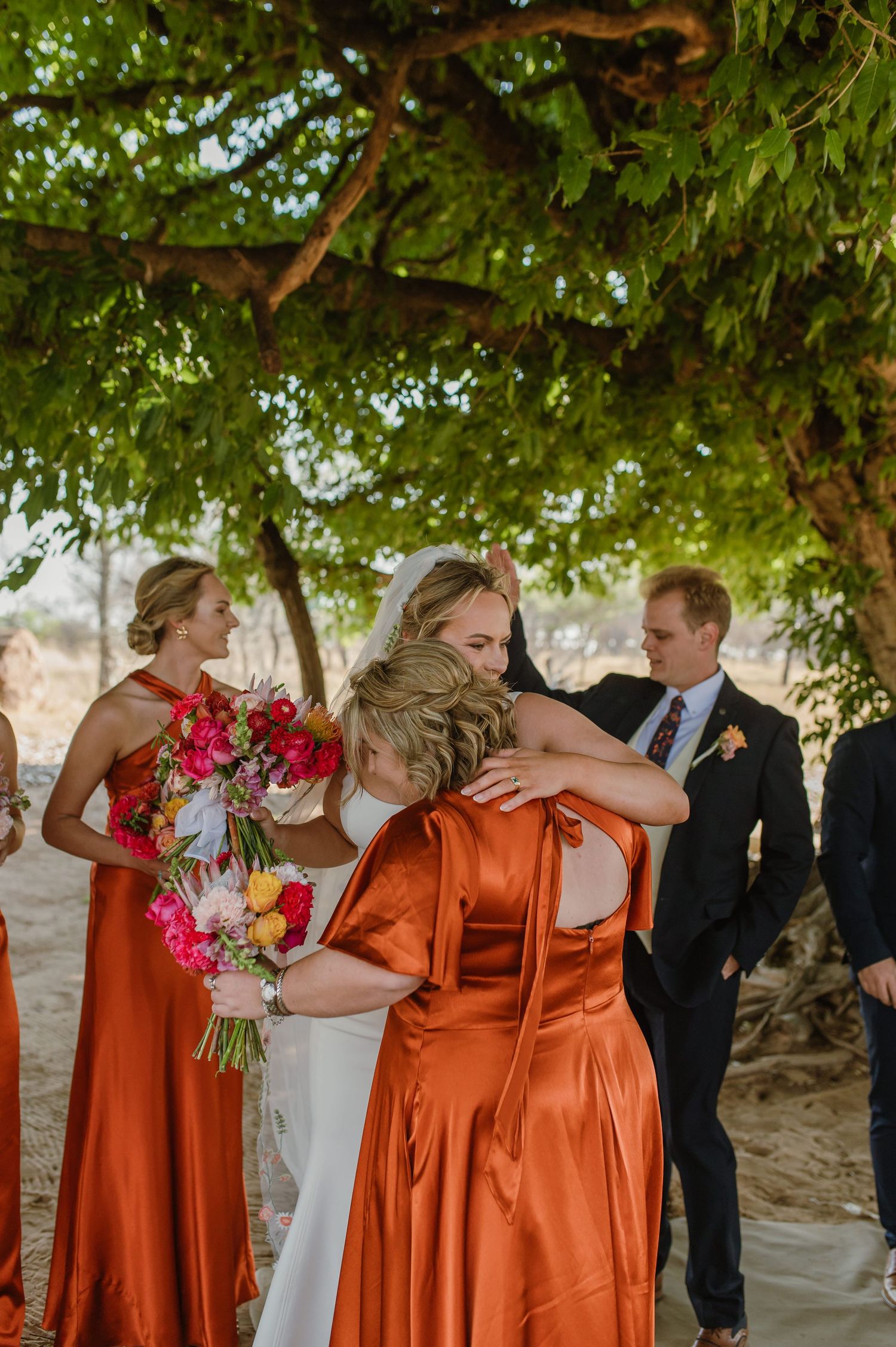 Wedding party in burnt orange dresses and dark suits celebrate under trees.