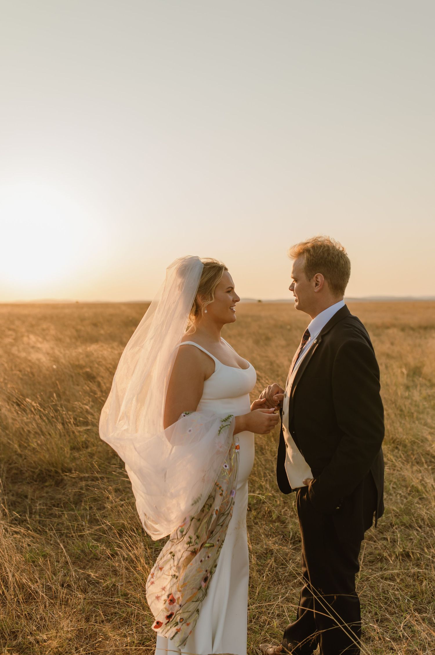 Newlyweds embracing in sunlit meadow with bride's veil flowing in wind.