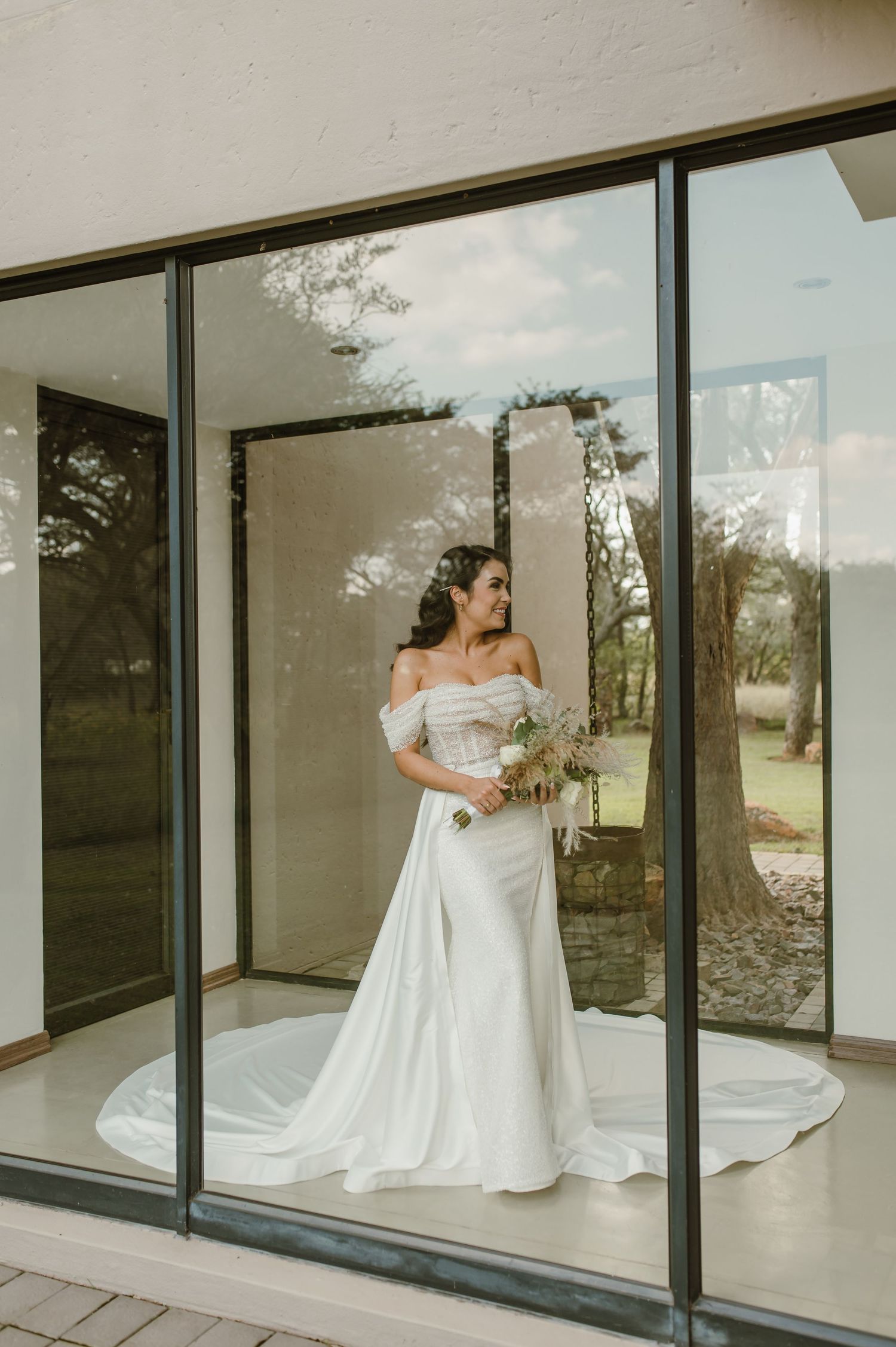 Wedding gown displayed against large glass windows in a contemporary white interior space.
