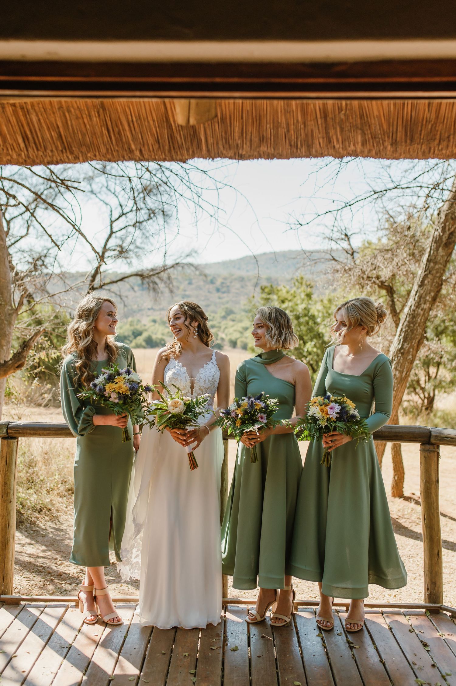 Wedding party lined up on wooden deck wearing sage green dresses with colorful bouquets.