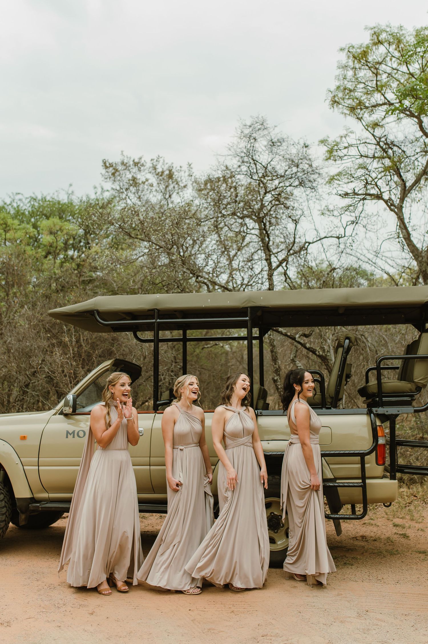 Bridesmaids in flowing taupe dresses pose excitedly by safari vehicle.