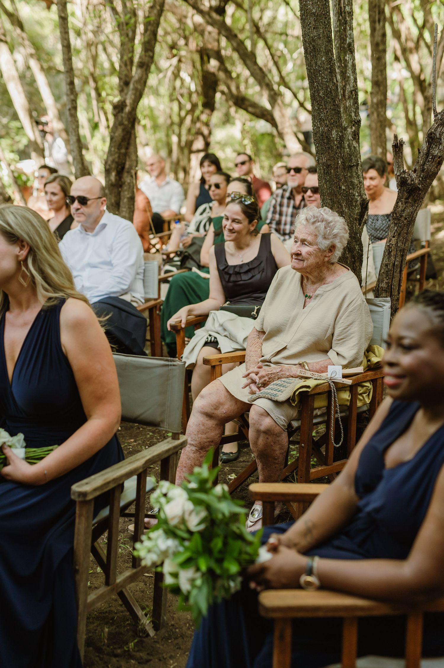 Wedding guests seated on wooden benches in a shaded outdoor forest venue during a ceremony.