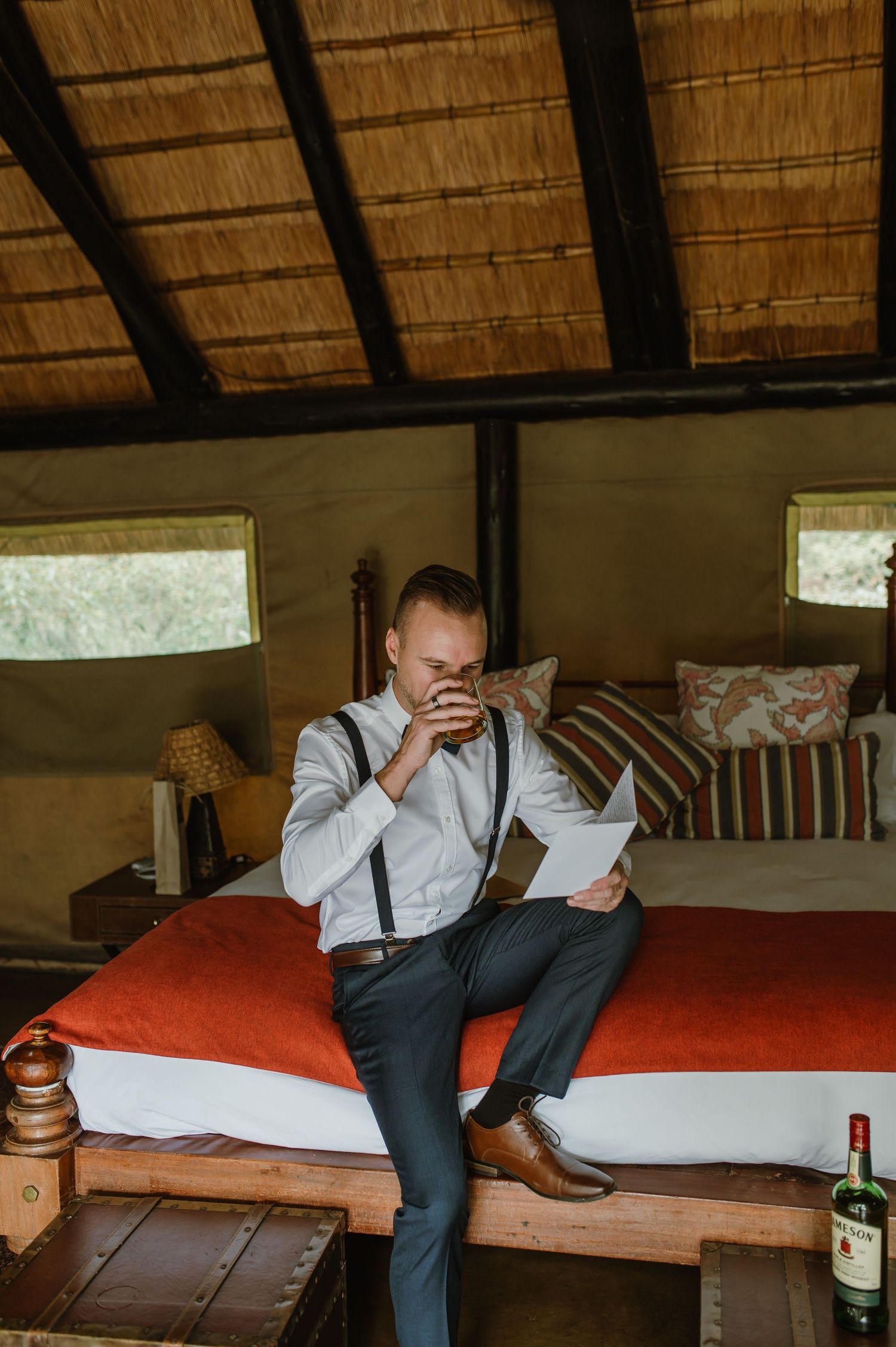 Someone in formal attire sitting on a bed in a rustic room with thatched roof.