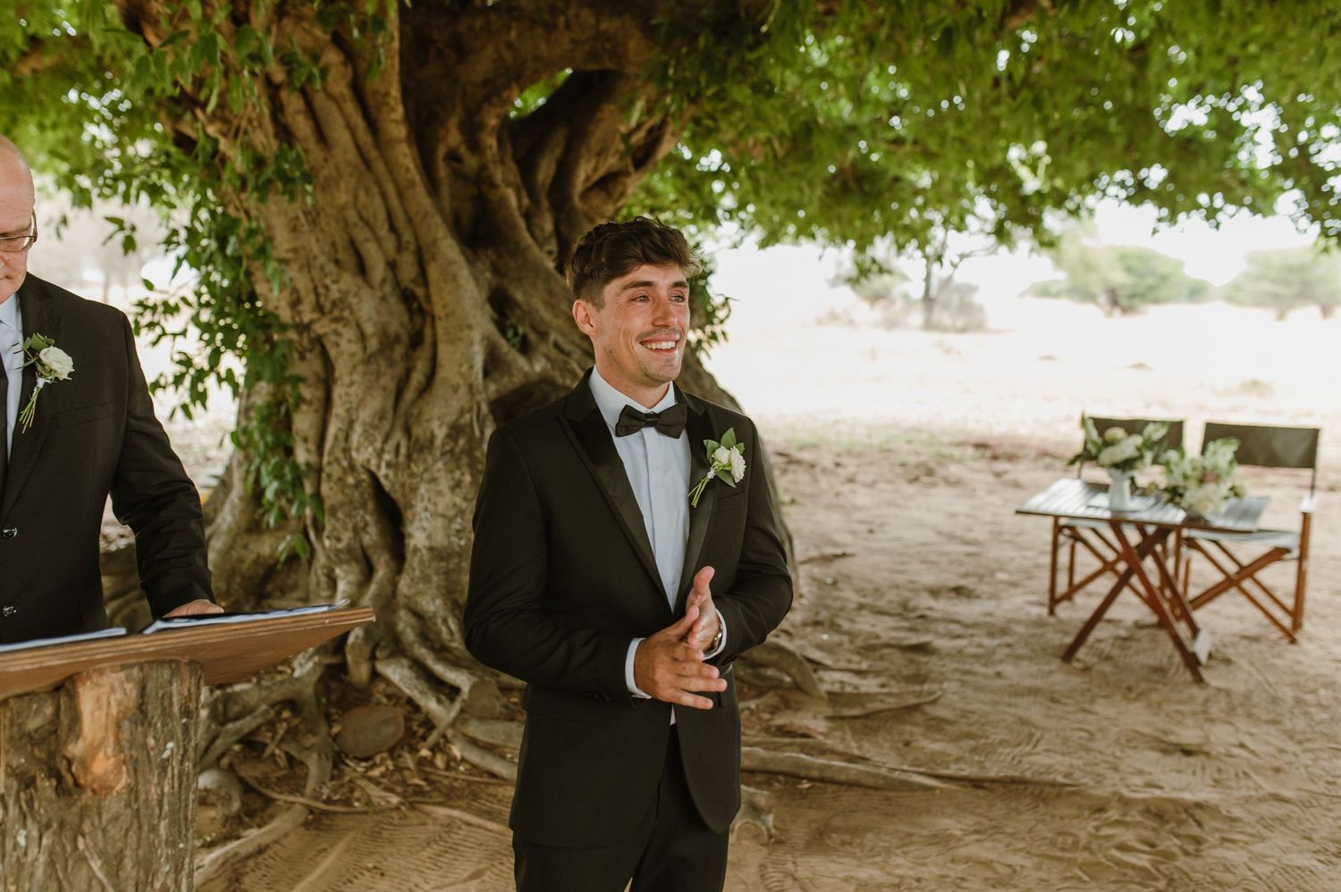 Outdoor wedding ceremony under trees with wooden benches and guests in formal attire.