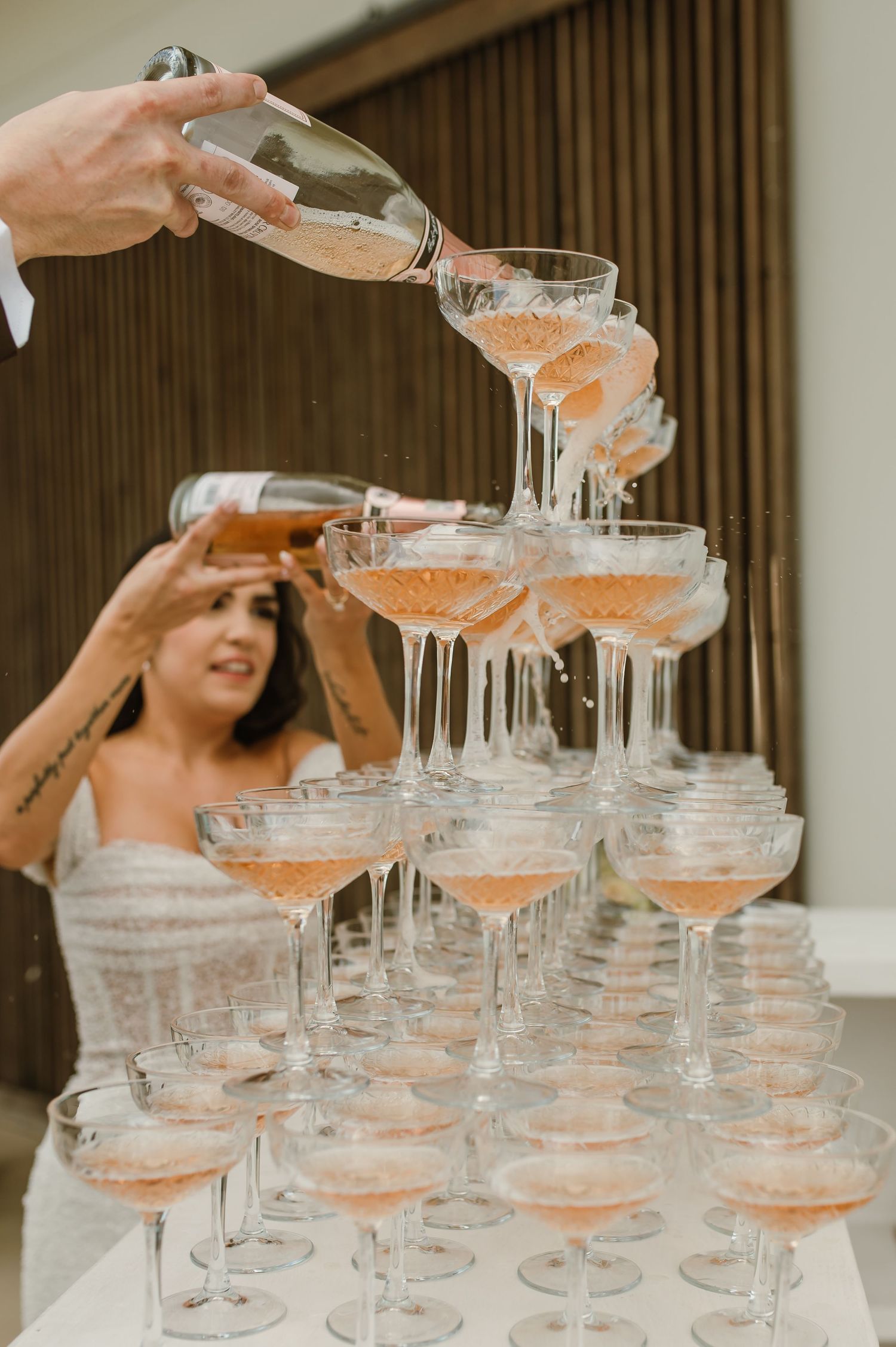 Hands pouring champagne into a tower of coupe glasses at an elegant wedding reception.