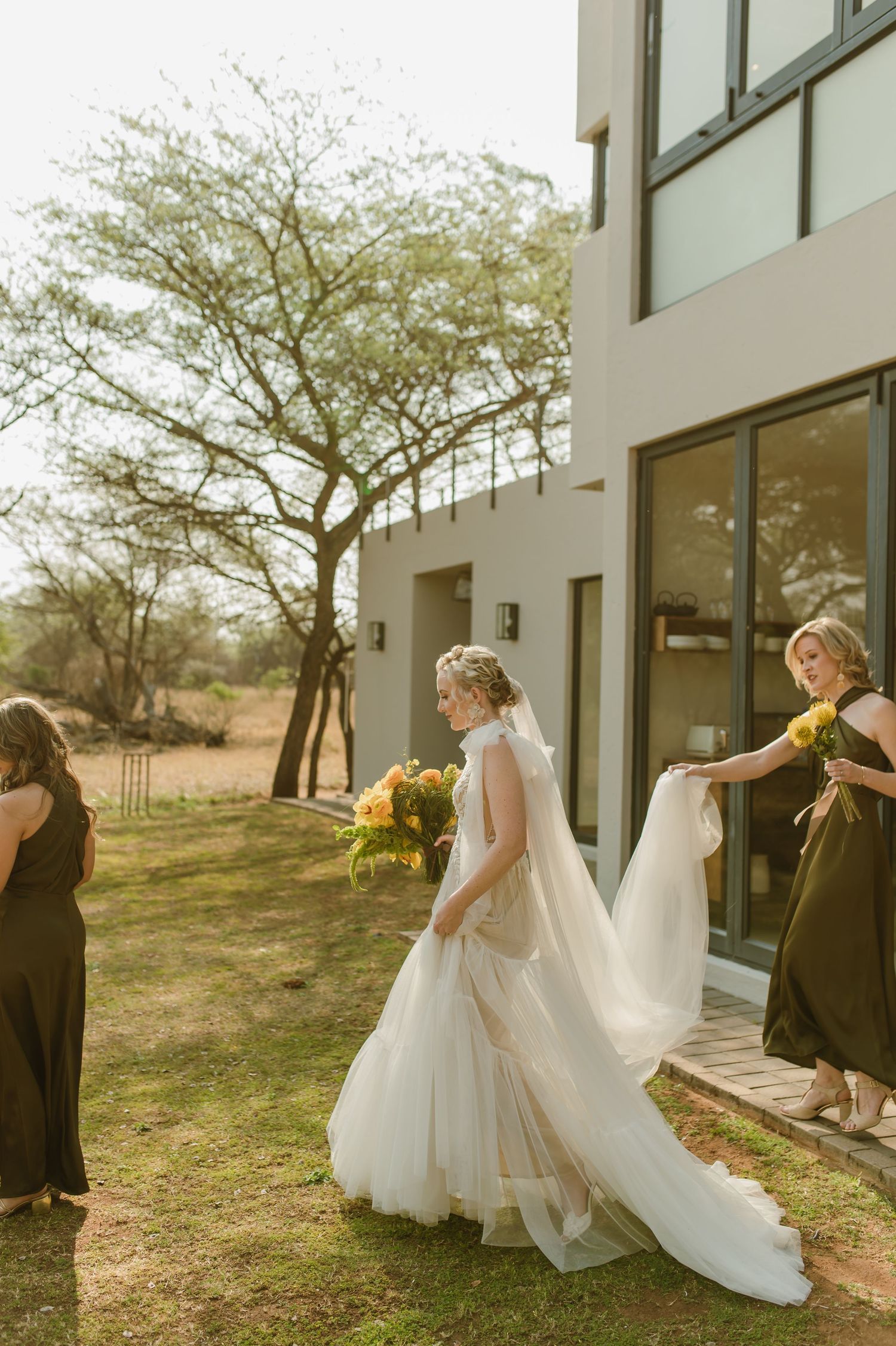Bridal party in white and black attire poses outside a modern building with sunlit trees.