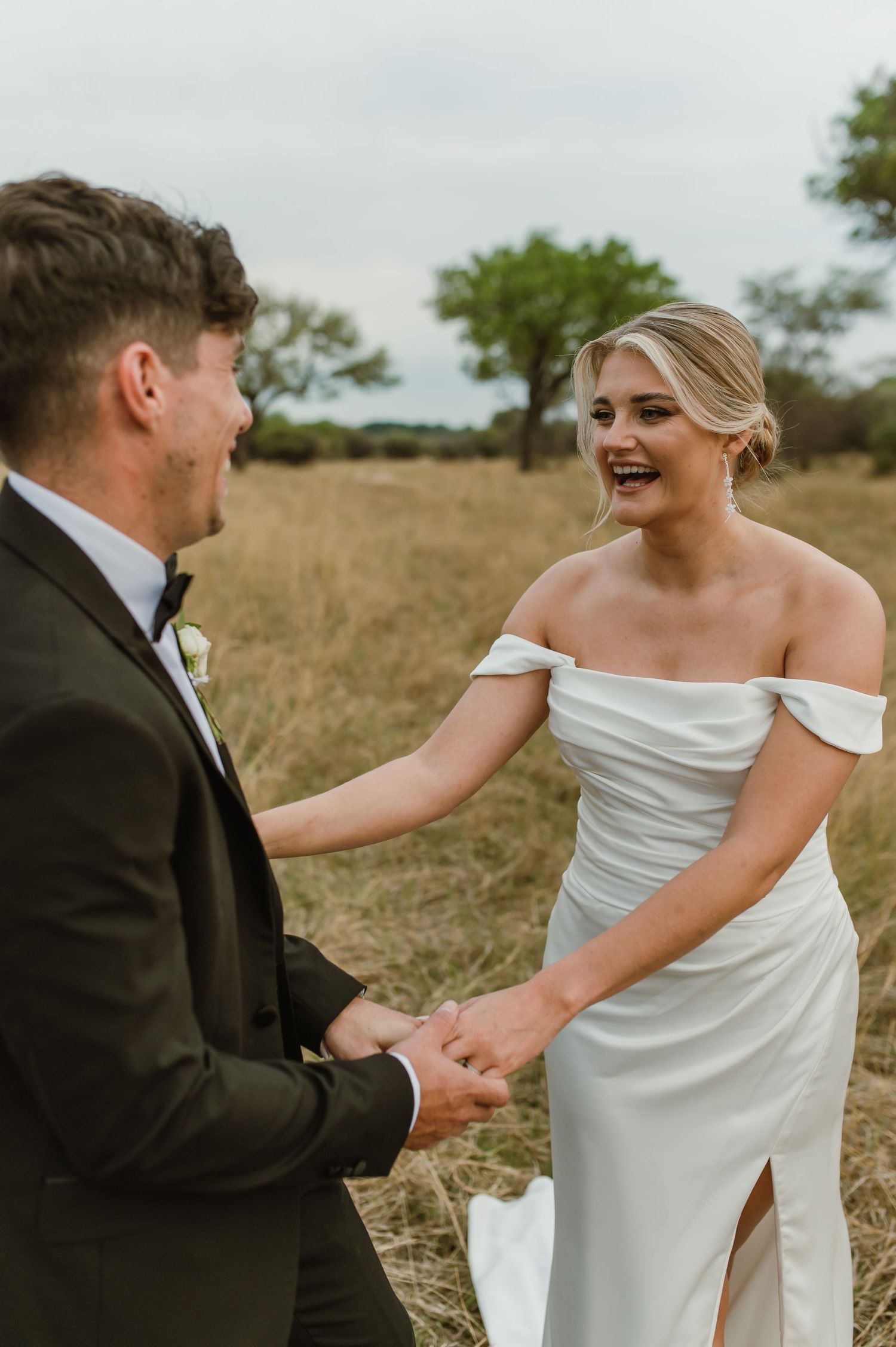 A bride in white dress and groom in tuxedo dance playfully in a grassy field.