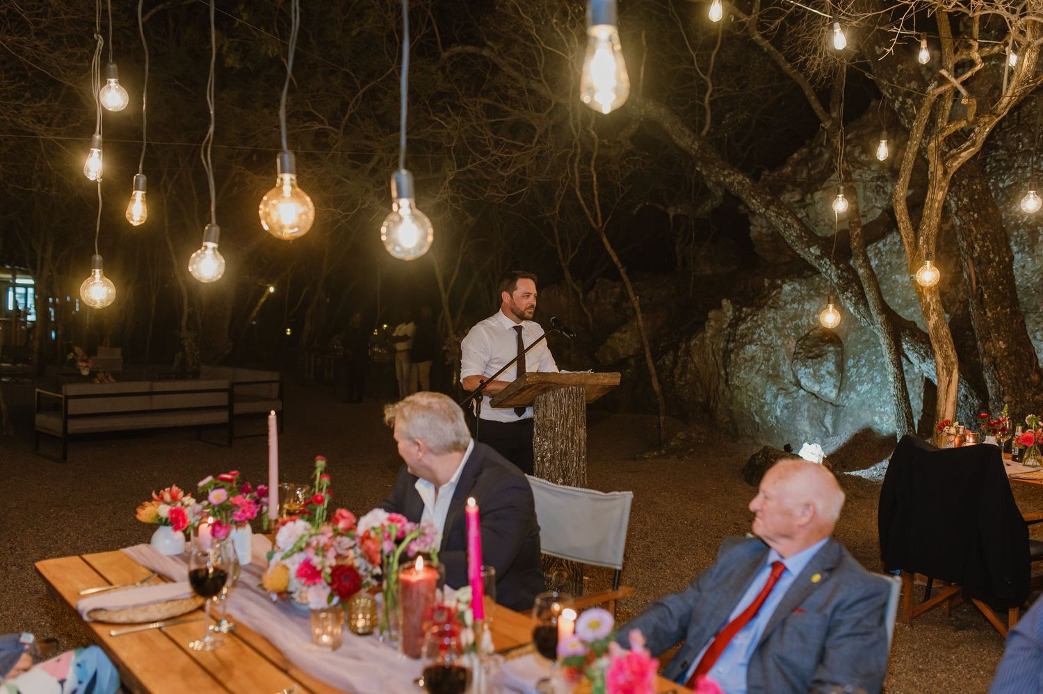 Nighttime outdoor celebration with guests seated at wooden tables under glowing string lights.