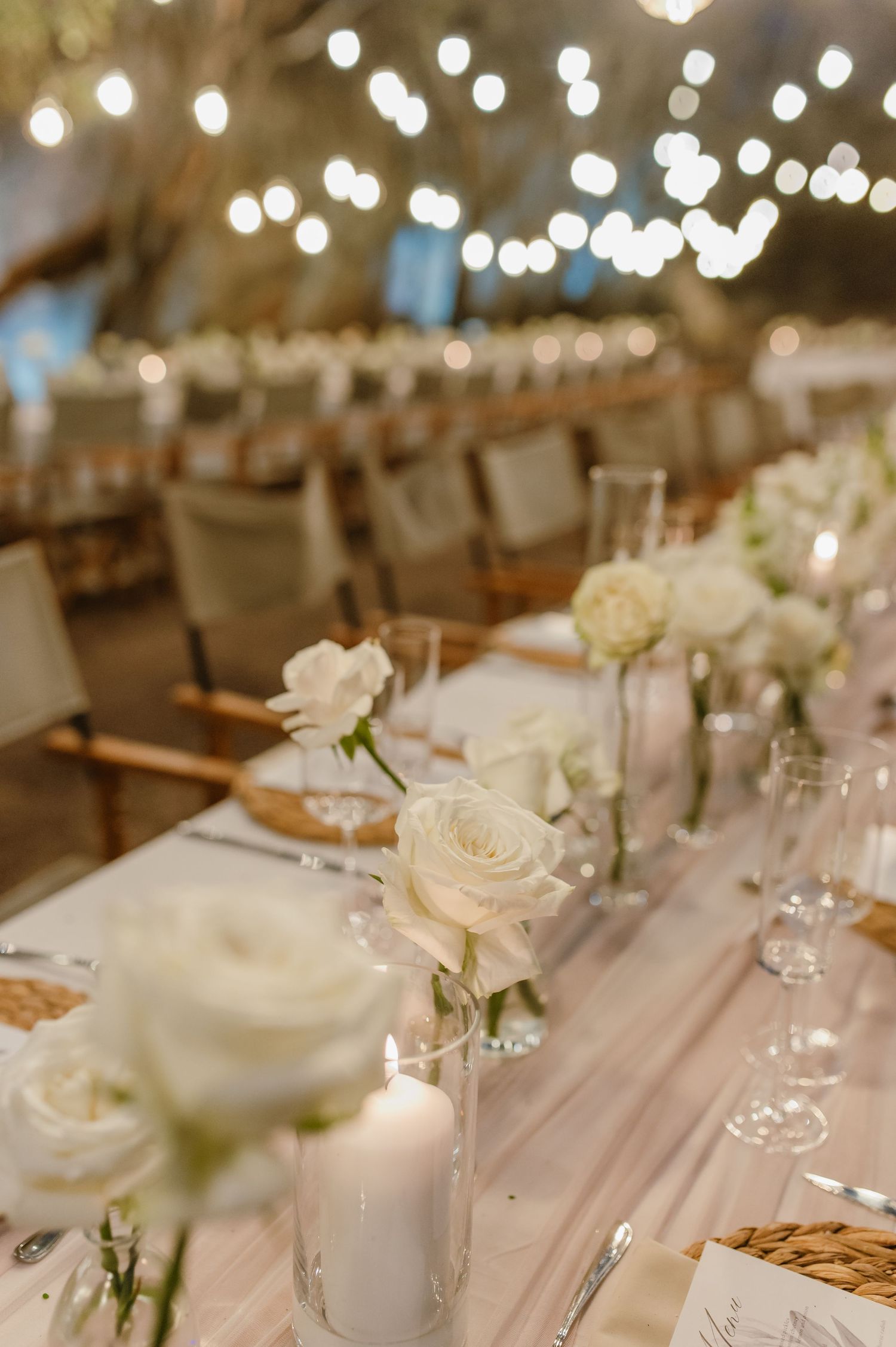 Elegant wedding reception table setting with white roses and string lights creating a romantic ambiance.