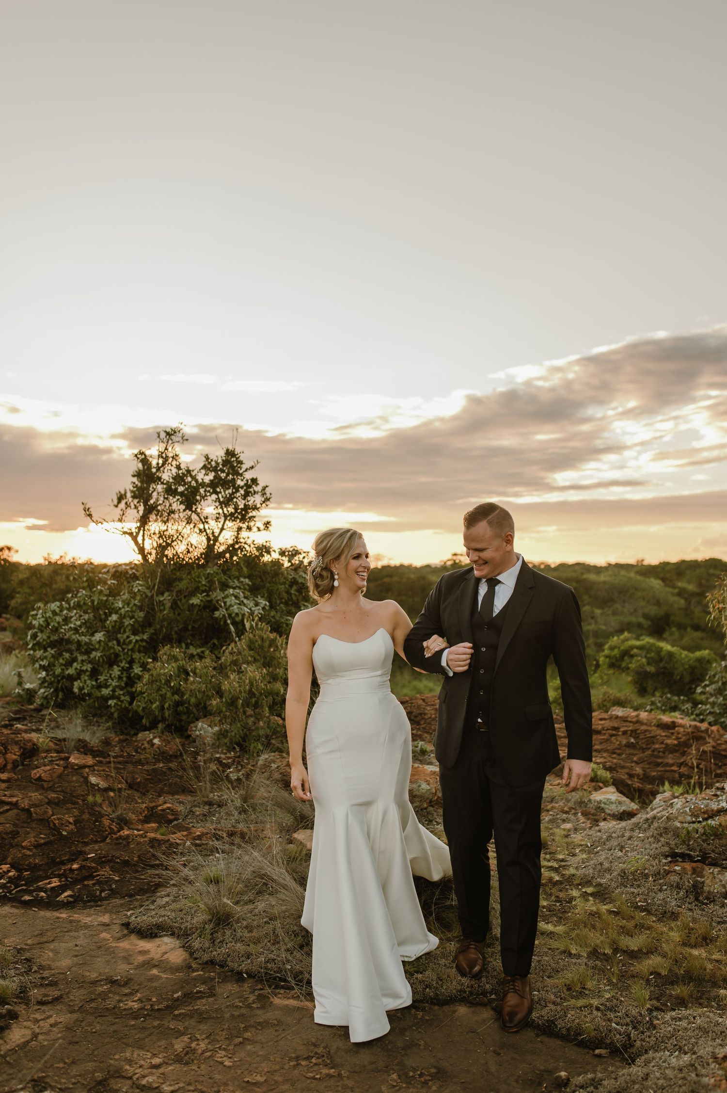 Wedding couple walks hand in hand through rugged desert terrain at sunset.
