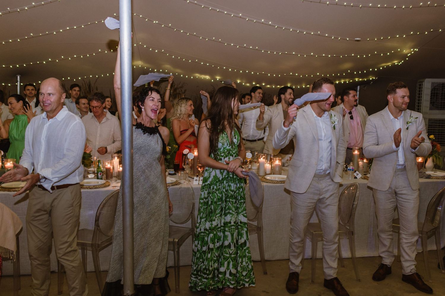 Wedding party members in khaki suits and colorful dresses dance together at an evening reception.