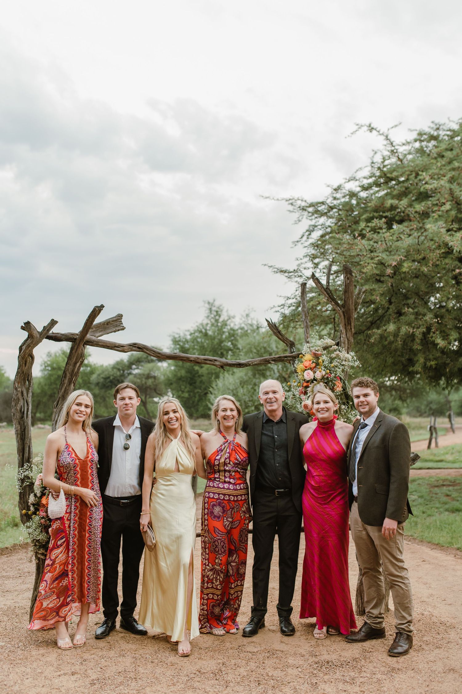 Wedding party members posed together outdoors wearing formal attire in red and neutral colors.