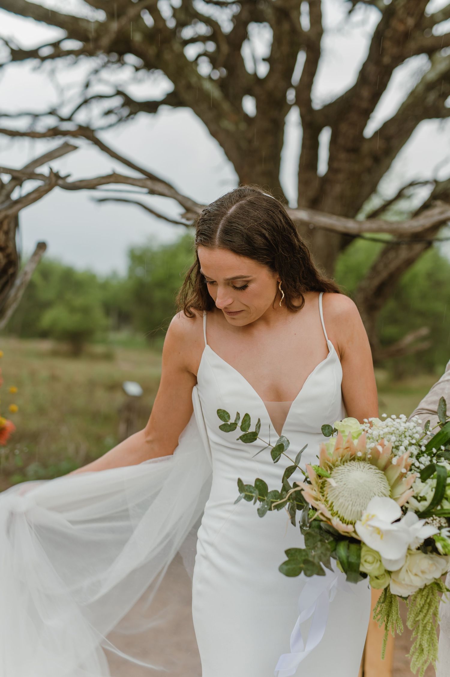 Elegant white wedding dress with flowing fabric and floral bouquet against a natural backdrop.
