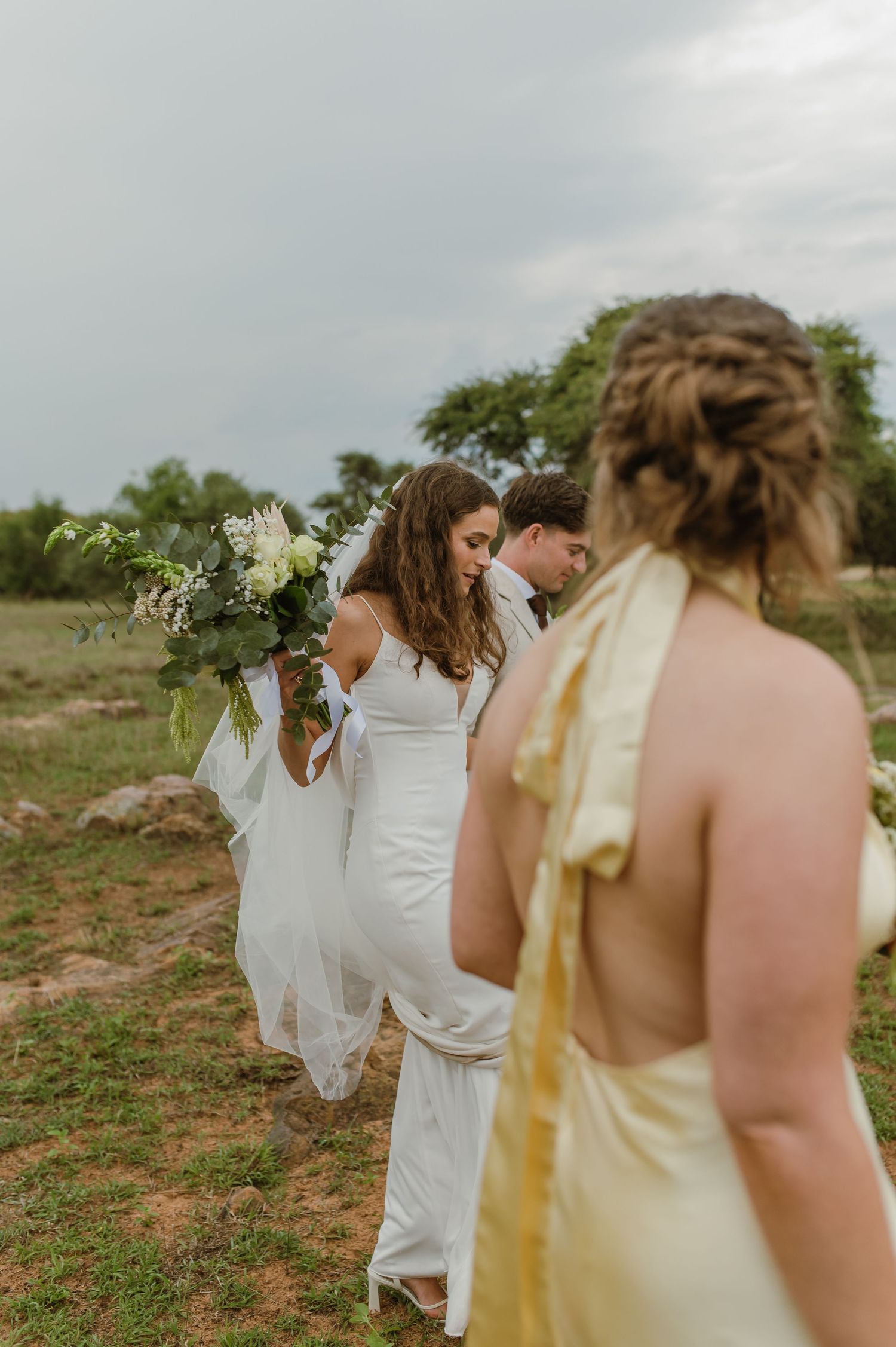 Wedding party members in white and yellow dresses gather outdoors against a cloudy sky.