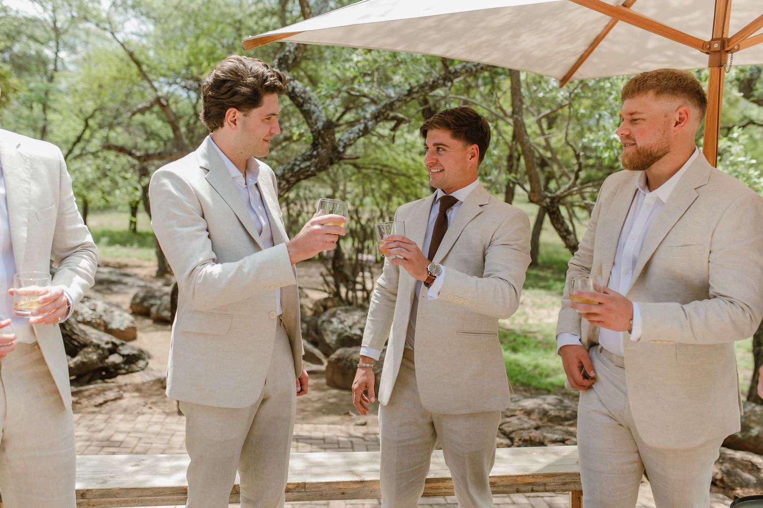 Groomsmen in light beige suits share laughter and drinks during an outdoor wedding celebration.