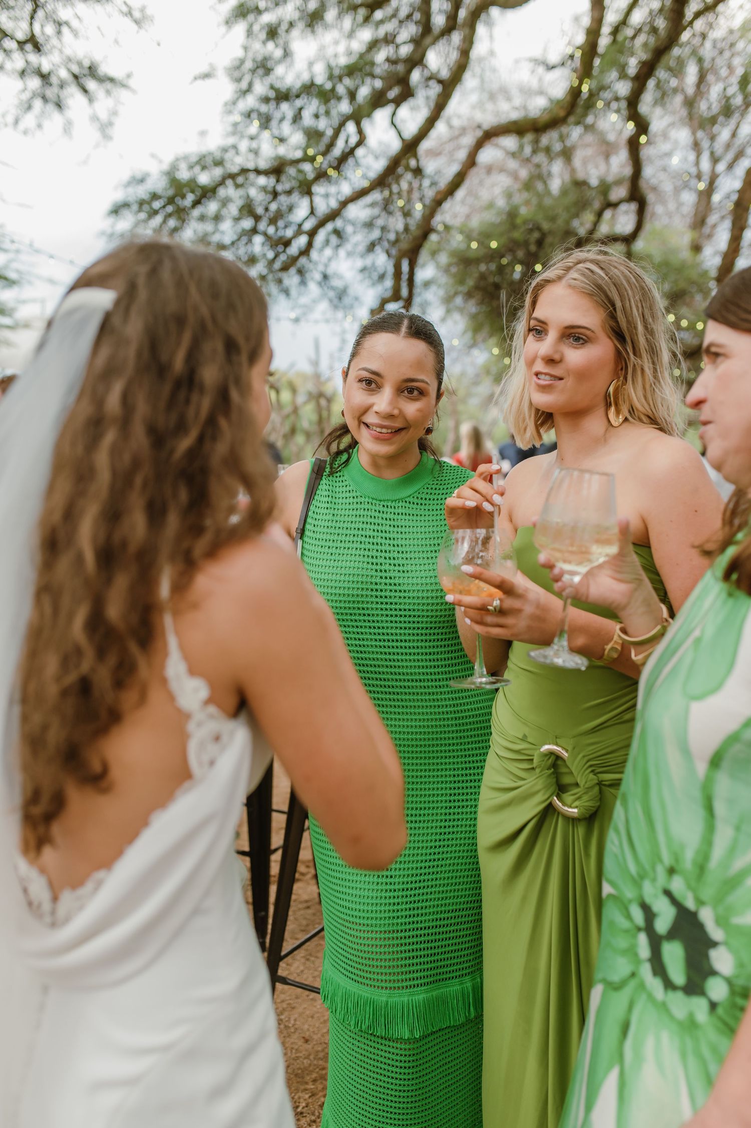 Bridesmaids in various shades of green dresses enjoying drinks at an outdoor celebration.