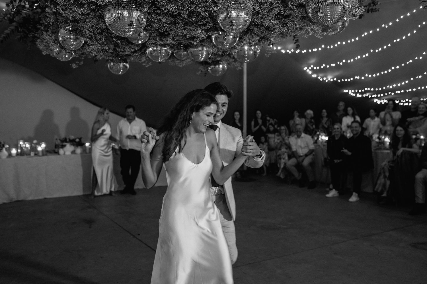 Black and white photo of a bride dancing solo under string lights and disco balls.