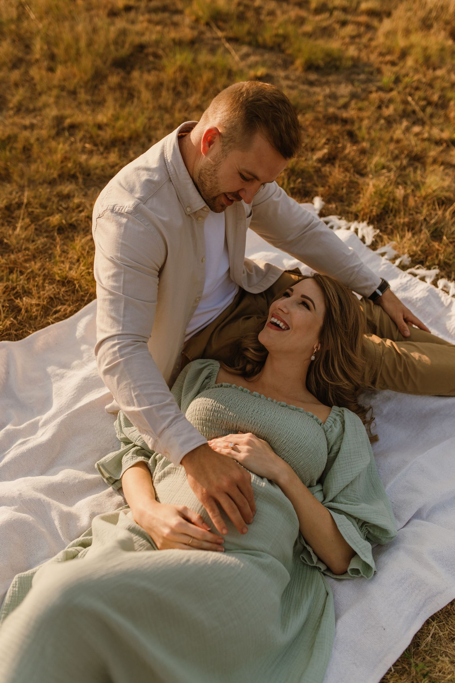 A couple in casual attire shares intimate moments while laying on a white blanket in a golden field at sunset.