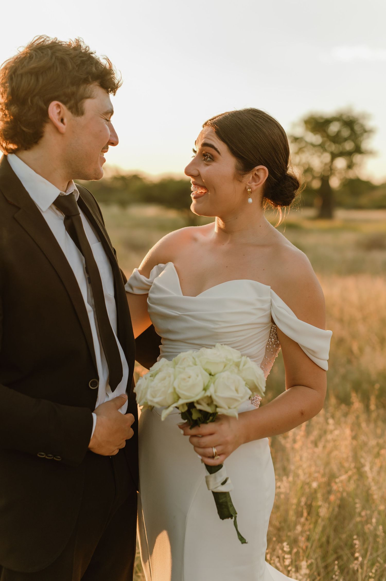 Bride and groom share a romantic golden hour moment in a field, bride holding white roses bouquet in off-shoulder gown.