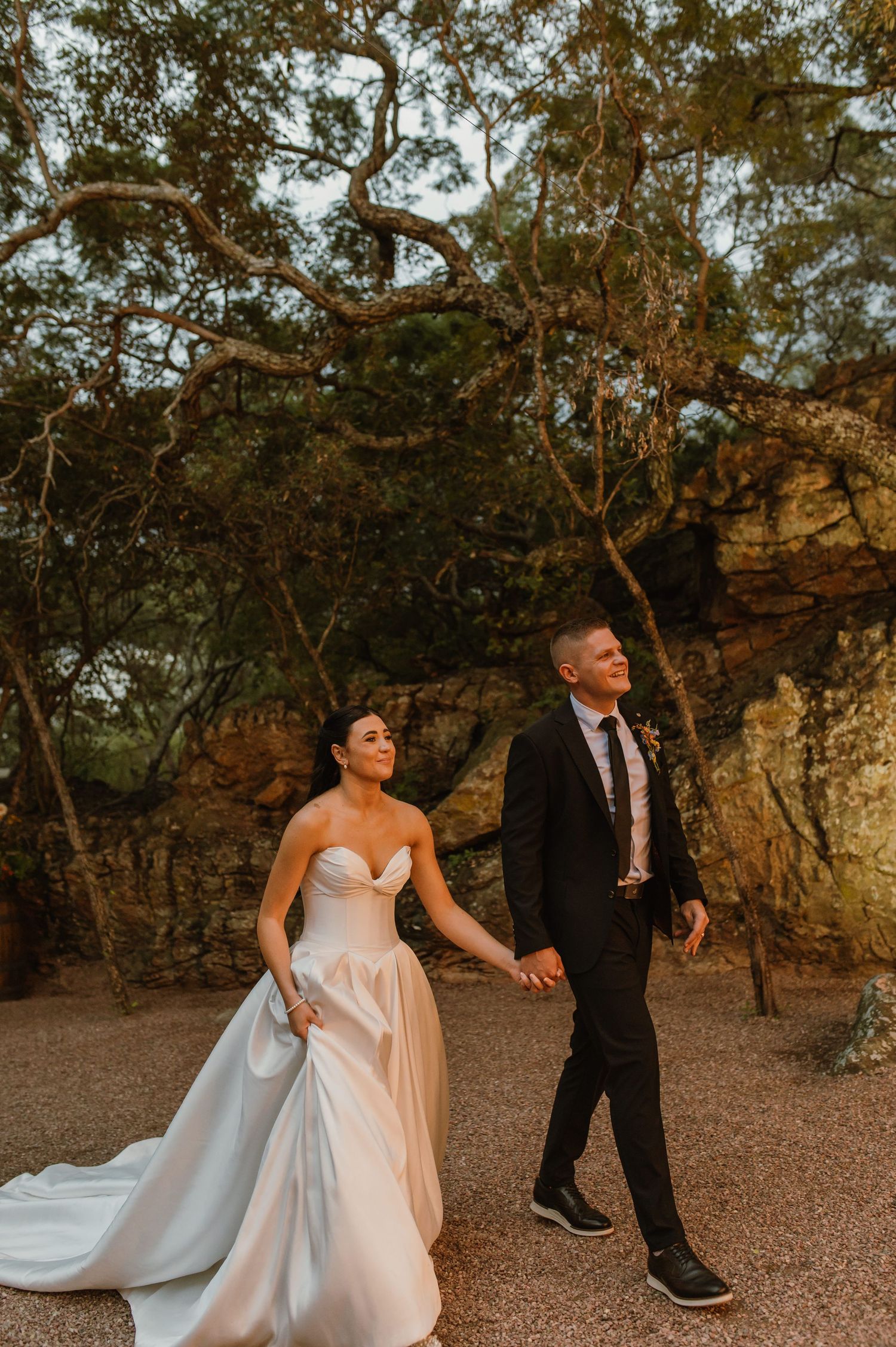 Newlyweds walking hand in hand through a scenic rocky tree-lined path in golden evening light.