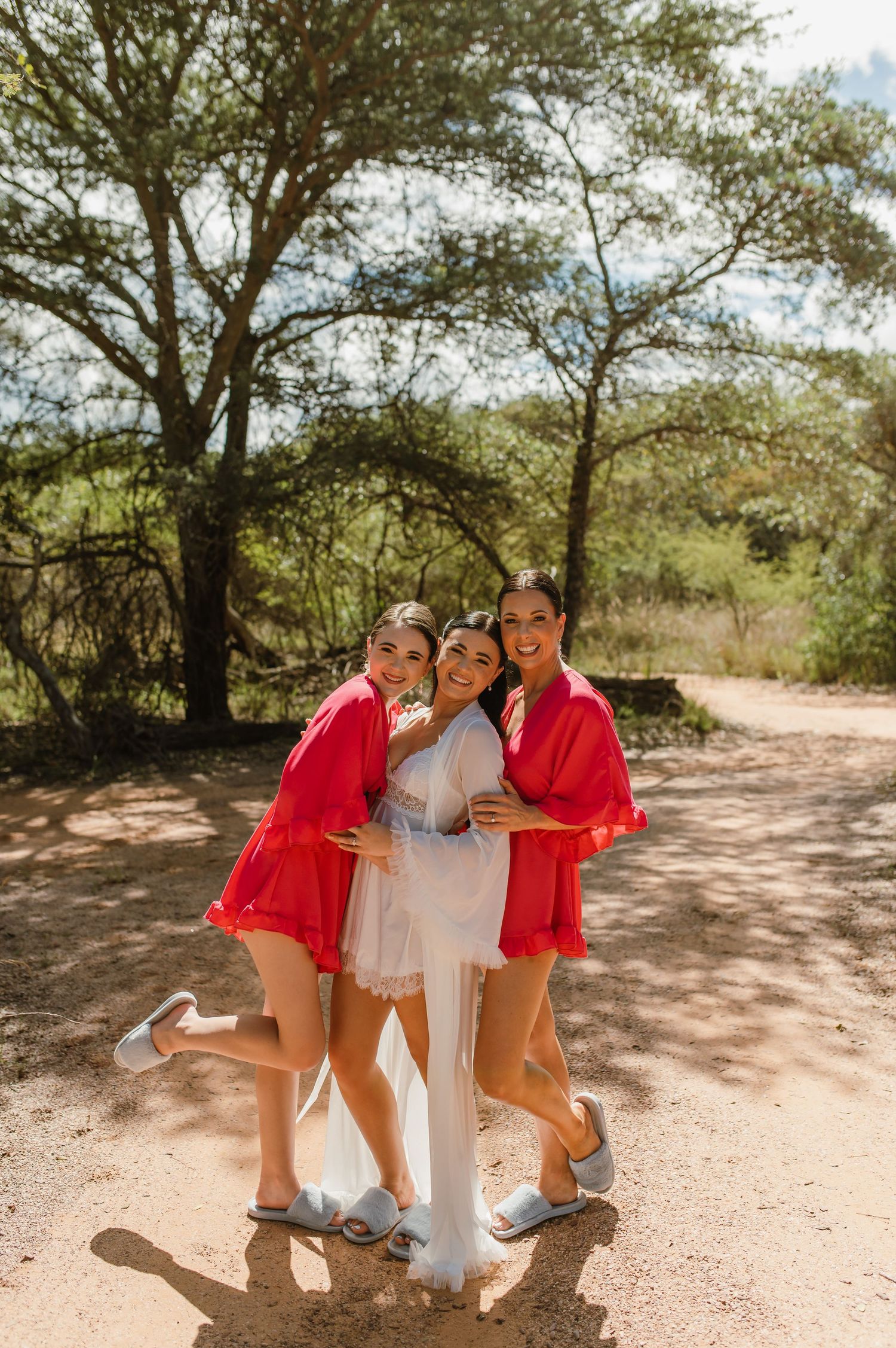 Bride in white robe with two bridesmaids in red robes hugging outdoors on a dirt path surrounded by trees.