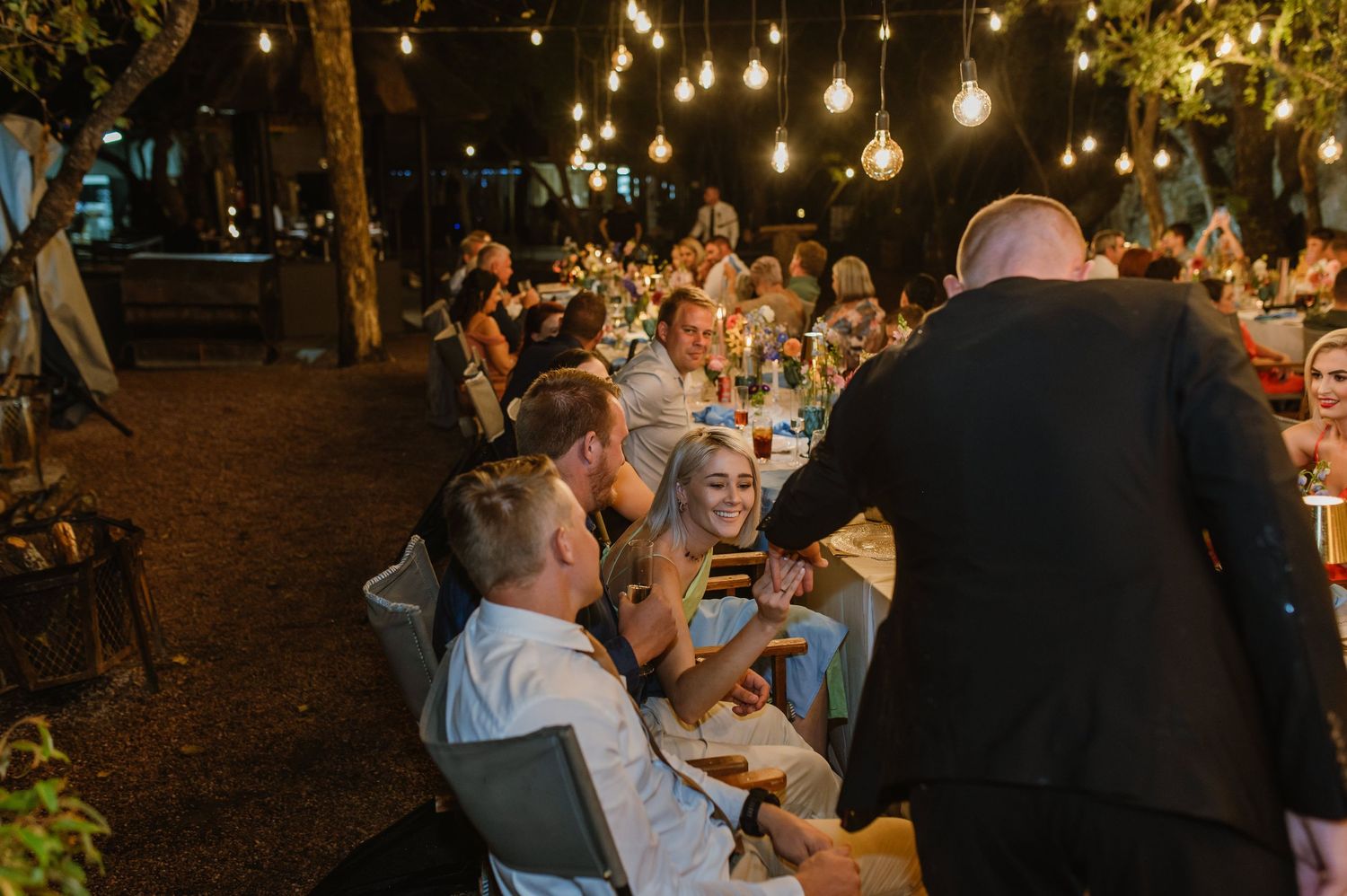 Joyful wedding guests laugh and celebrate at candlelit outdoor evening reception under string lights.