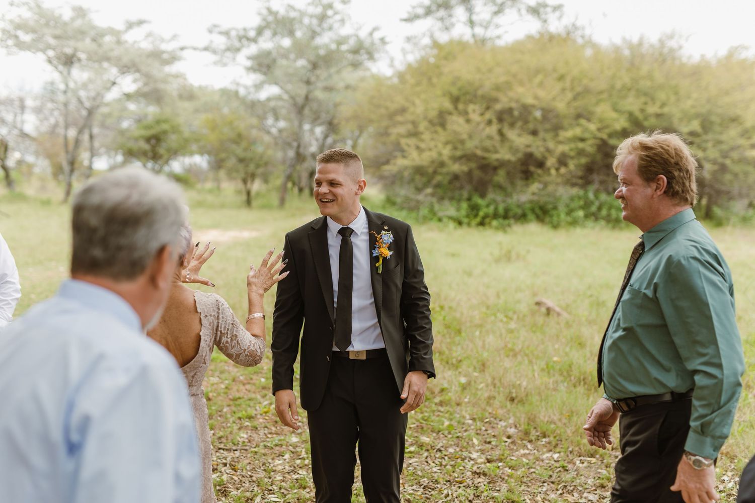 Groom in black suit laughing with guests in a lush green outdoor setting before the ceremony.