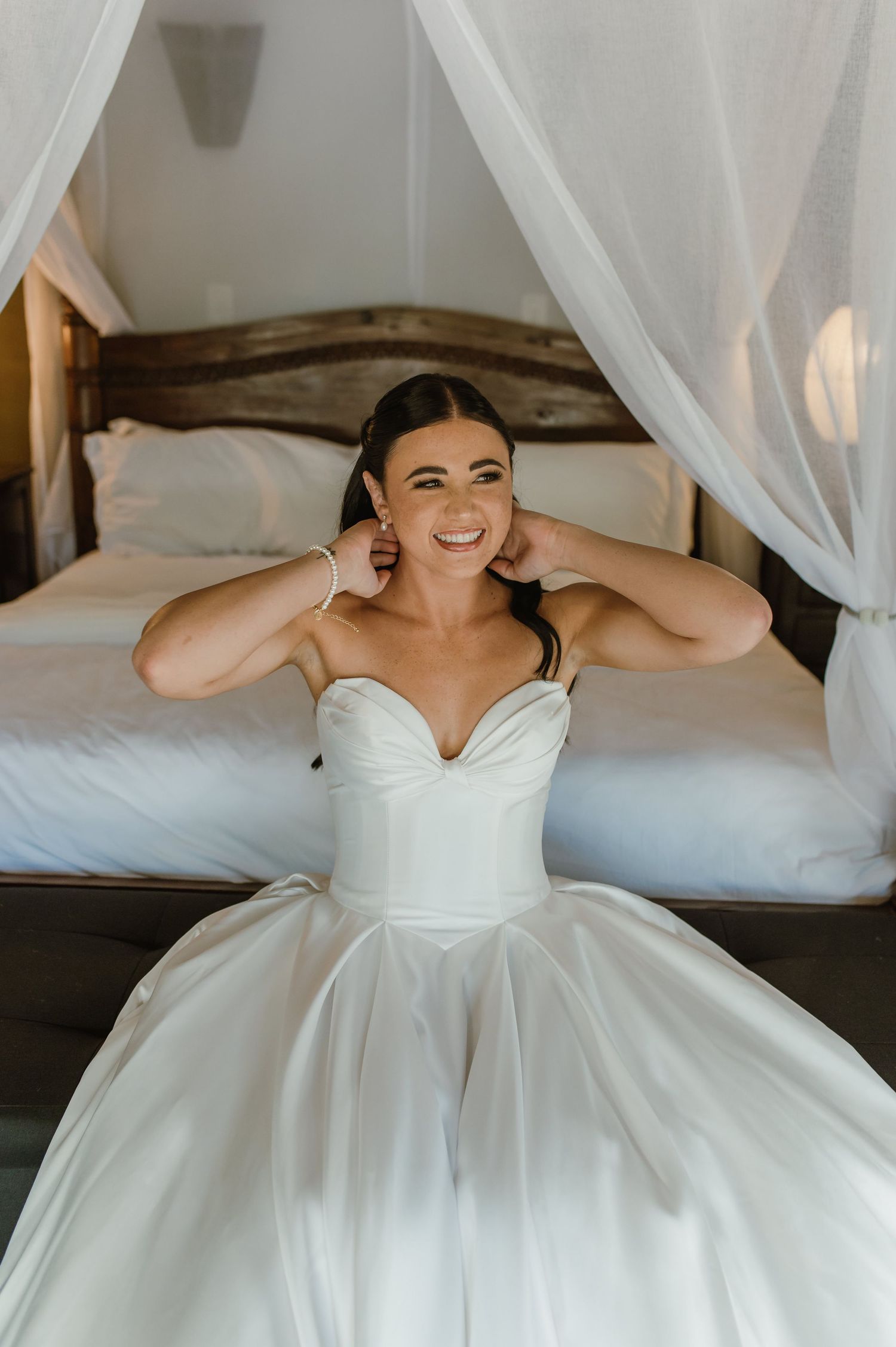 Smiling bride in strapless ballgown putting on earrings beside a canopy bed in a lodge room.
