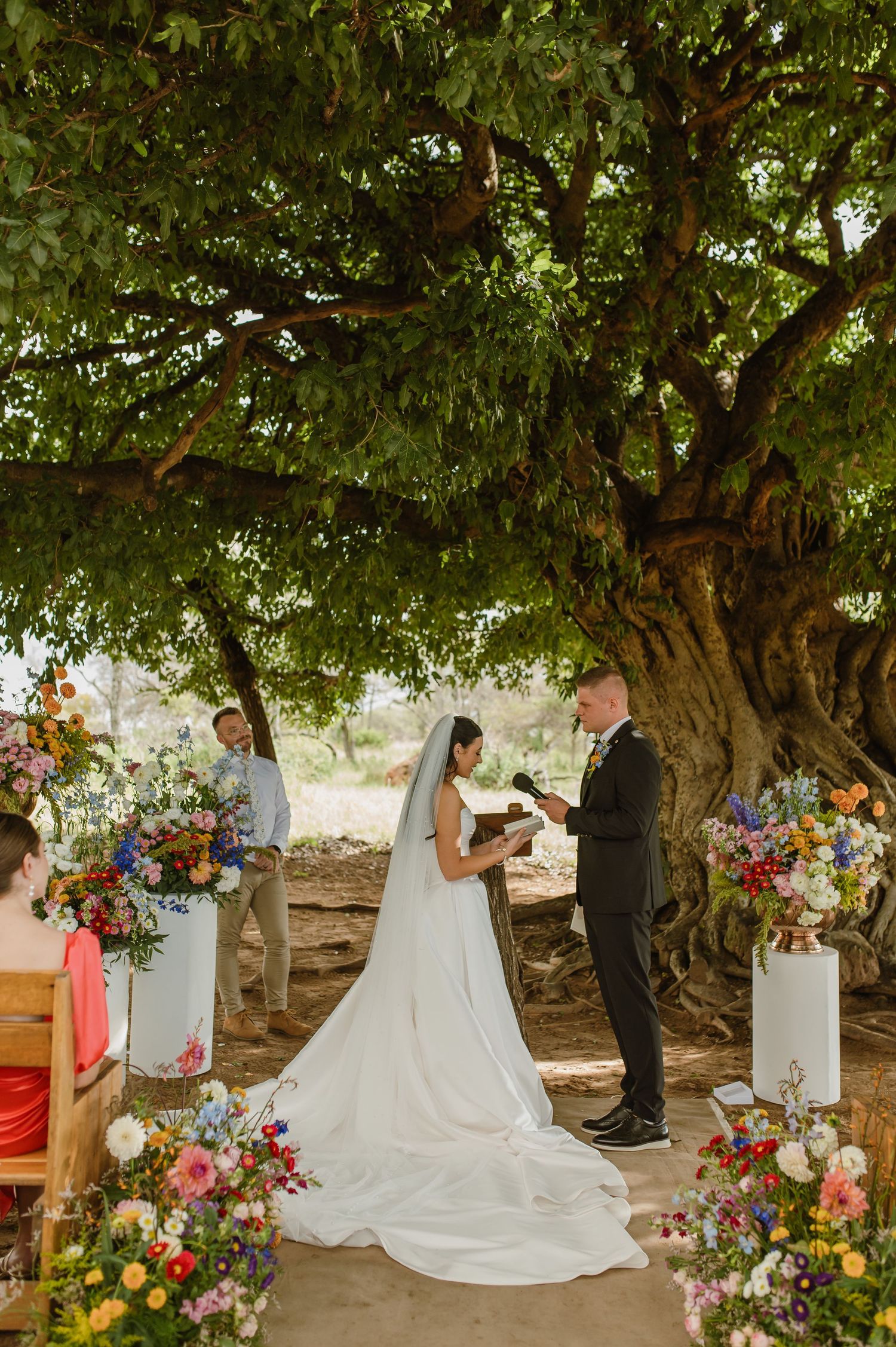 Bride and groom exchanging vows under a grand tree flanked by vibrant floral arrangements at an outdoor wedding.