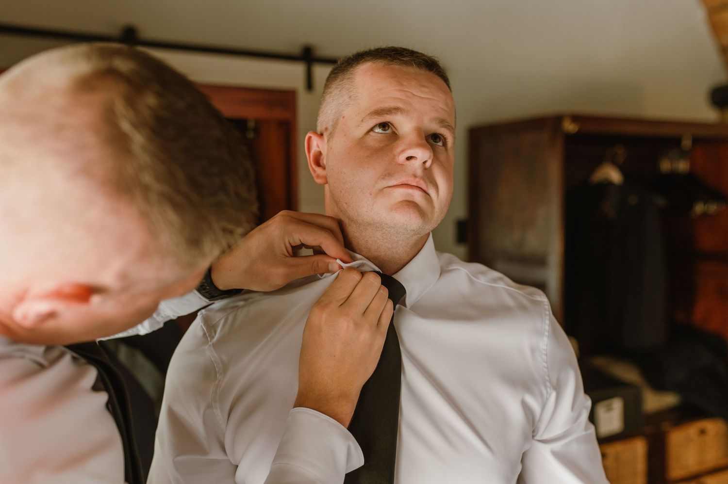 Groomsman adjusting the tie of a groom in a white dress shirt during wedding preparations indoors.