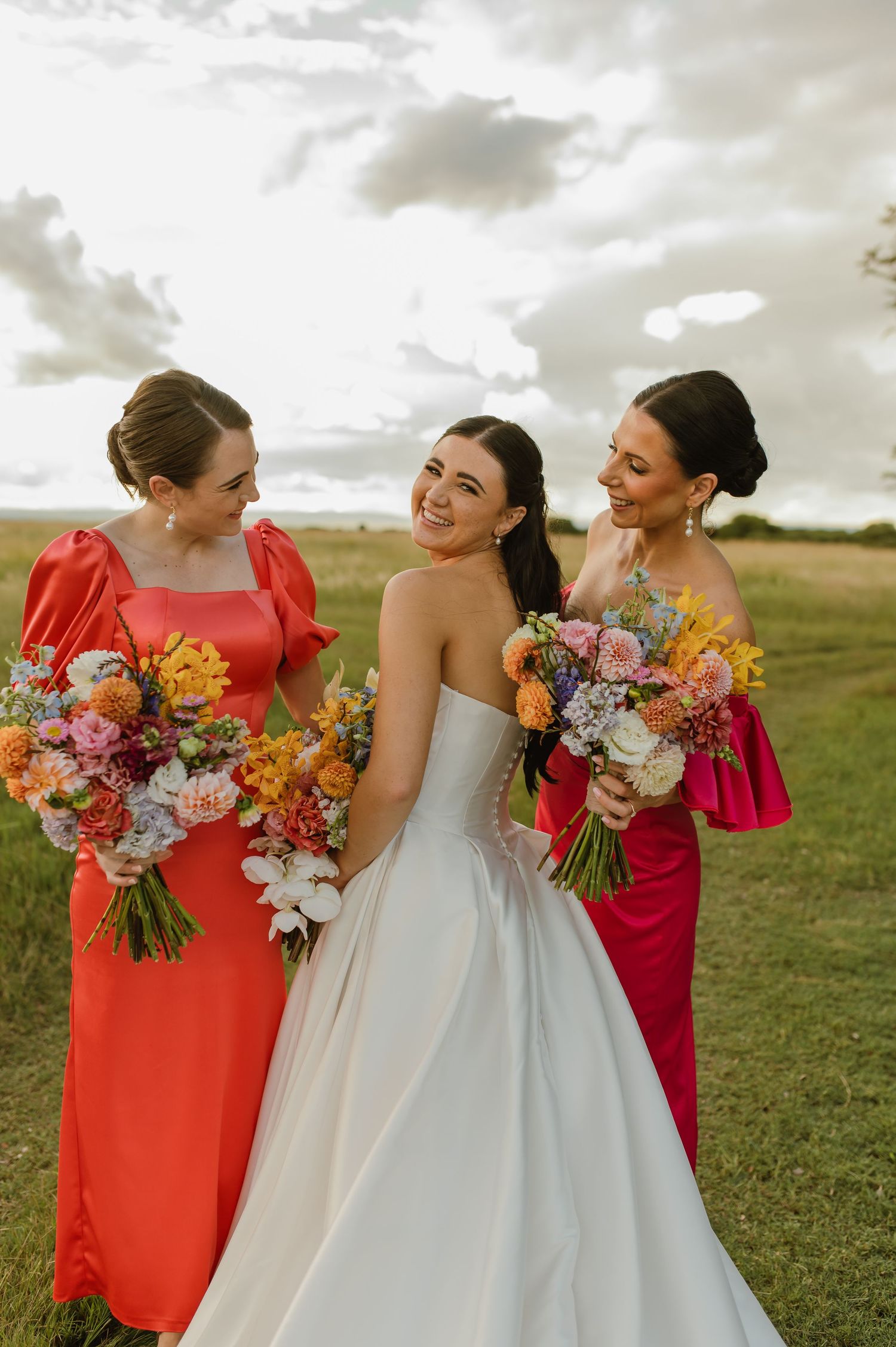 Bride in strapless white gown poses with two bridesmaids in red dresses holding colorful bouquets outdoors.