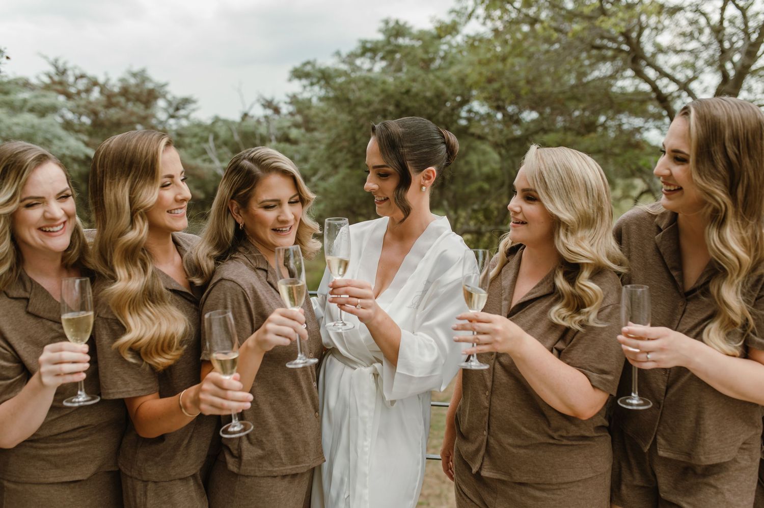 A group of friends in matching brown outfits and one in white share champagne glasses while celebrating outdoors.