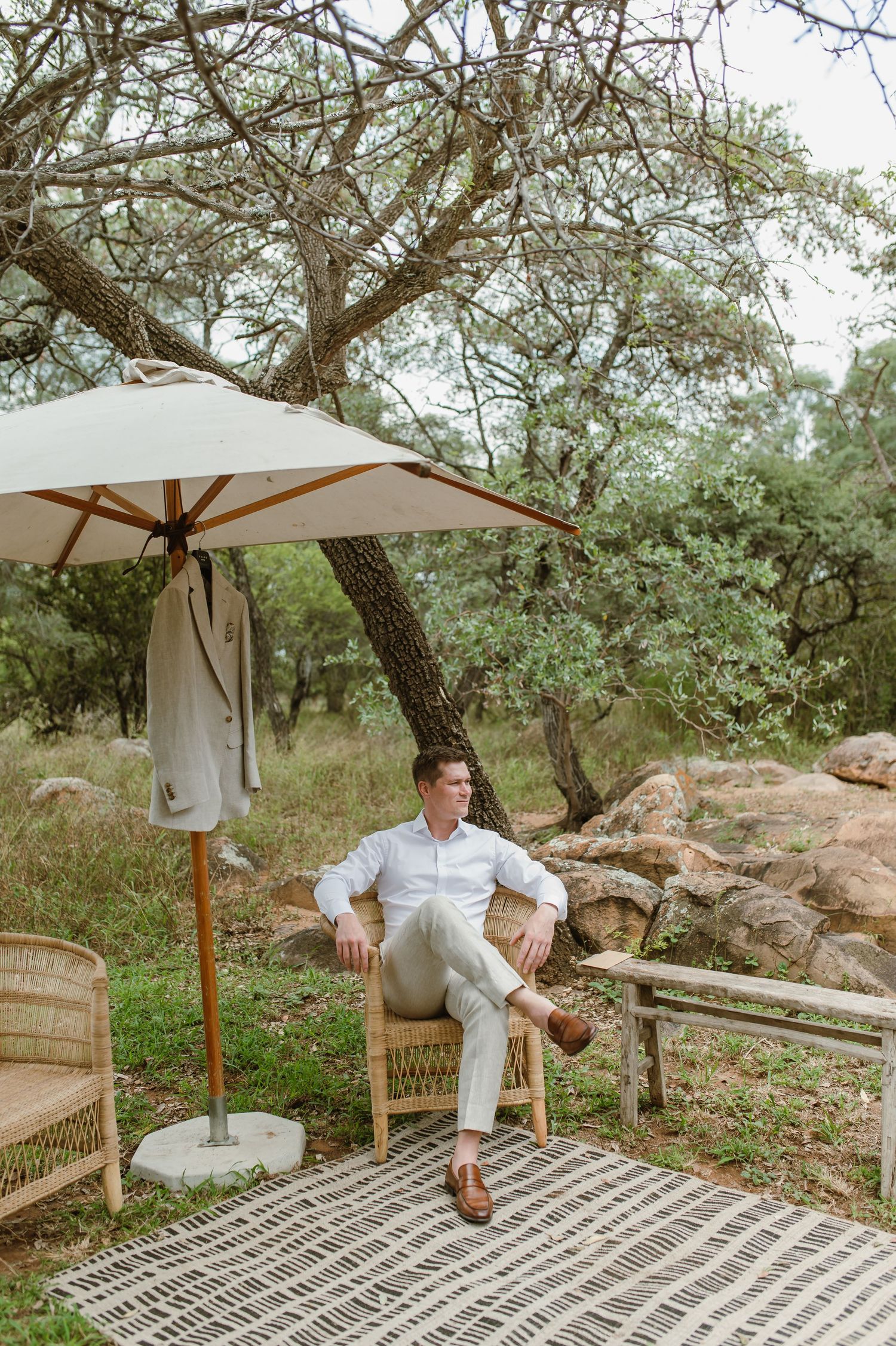 A relaxed outdoor seating area with a wicker chair and white umbrella on a patterned rug surrounded by trees and rocks.