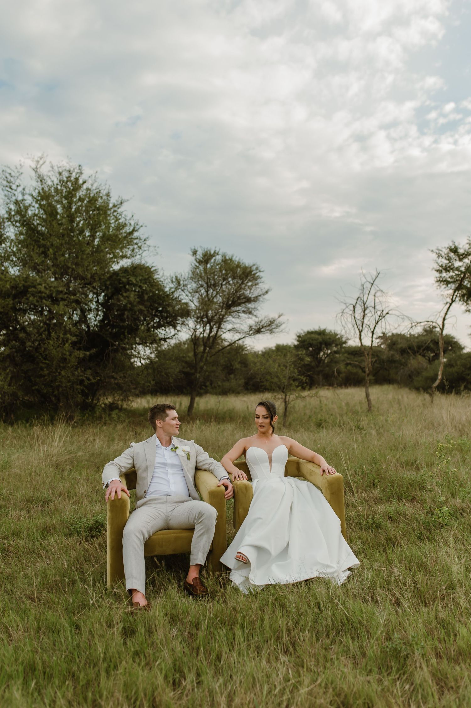 A couple in wedding attire sits together on a yellow couch in an open field during a sunny day.