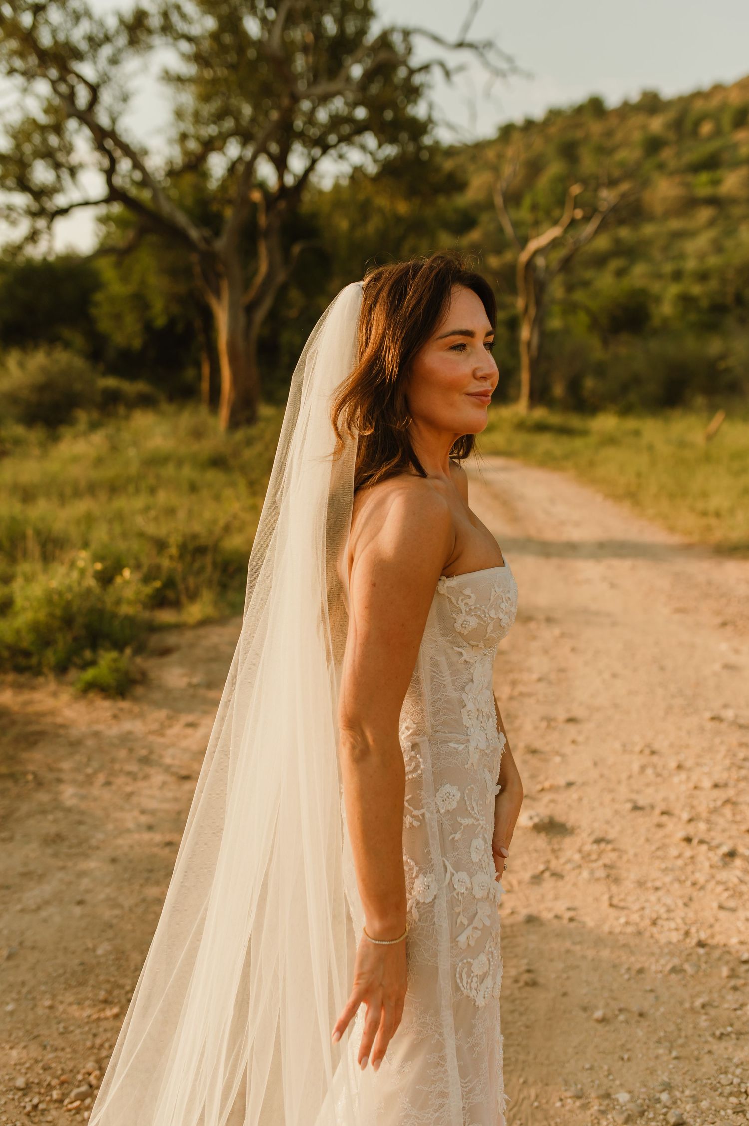Bride in strapless lace wedding gown and flowing veil stands on dirt path surrounded by lush greenery at golden hour.