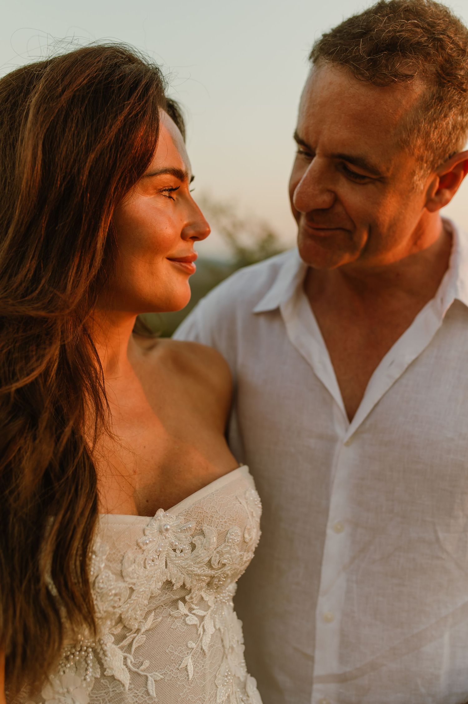 Bride and groom share an intimate glance during golden hour, bride in lace gown, groom in white linen.