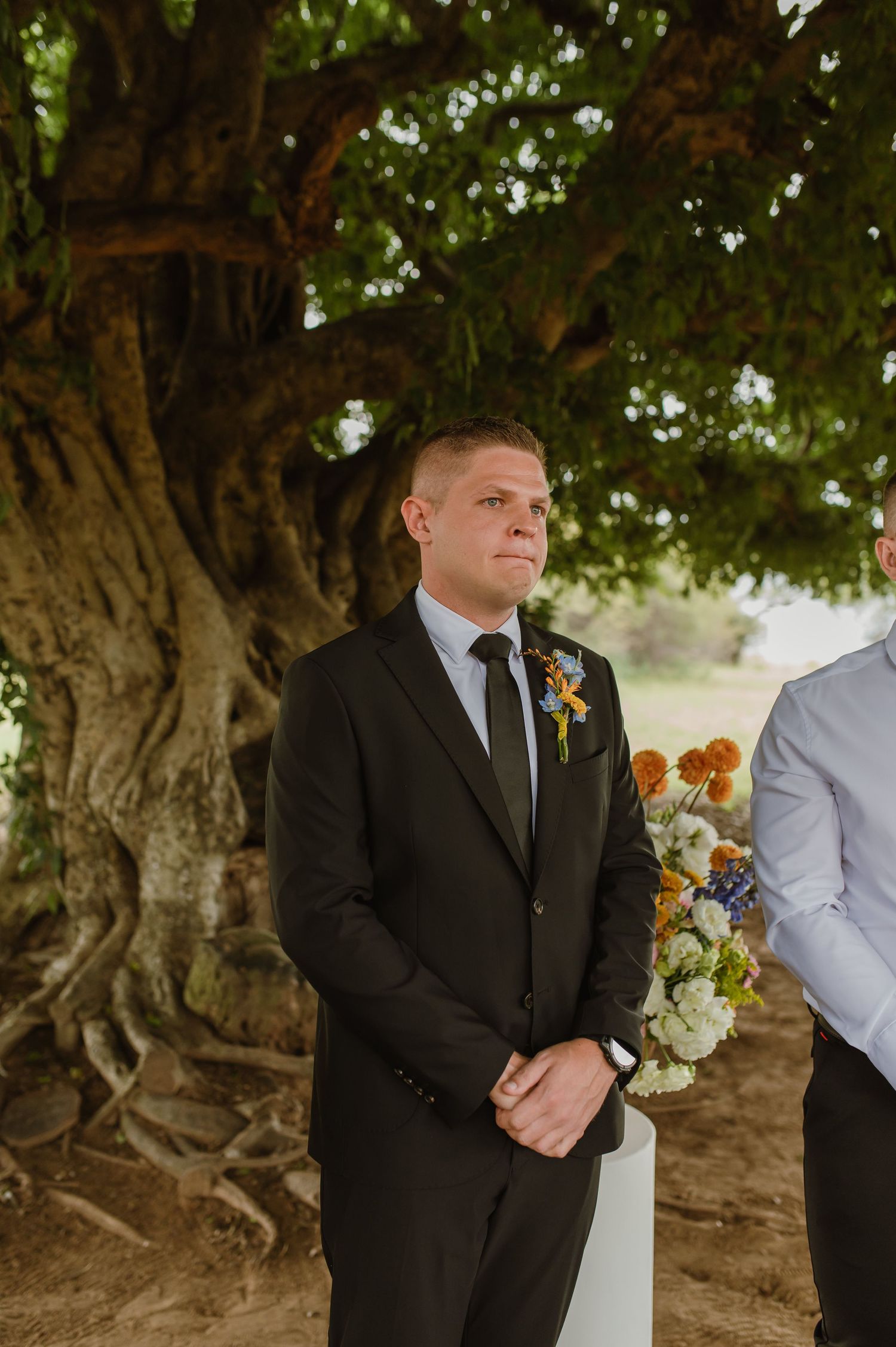 Groom in black suit with floral boutonniere standing at the altar beneath a large tree awaiting his bride.