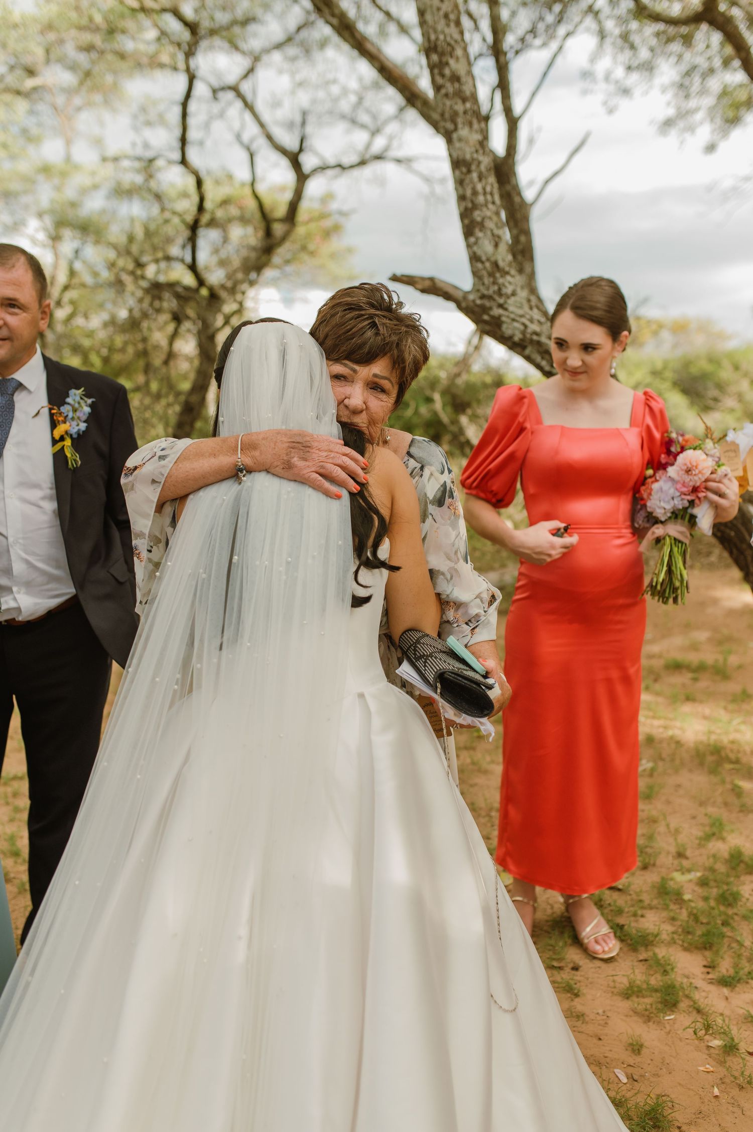 Emotional mother hugging bride in white wedding dress outdoors while bridesmaid in red watches.