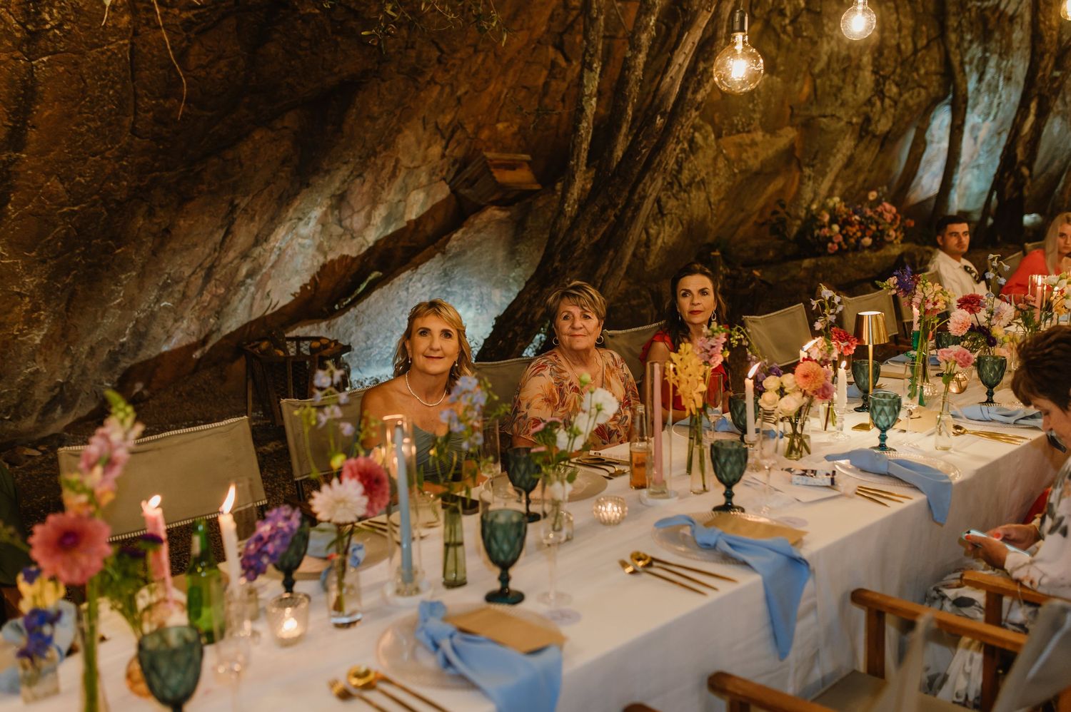 Wedding reception guests seated at elegantly decorated table with colorful flowers and candles in cave venue.