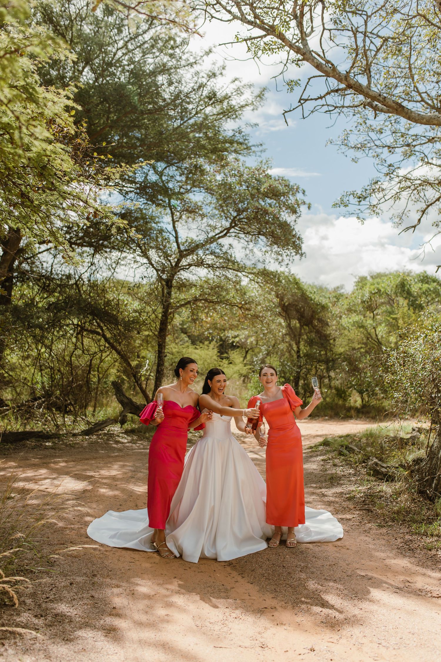 Bride in white gown toasting with two bridesmaids in red and orange dresses on a sandy bush path.