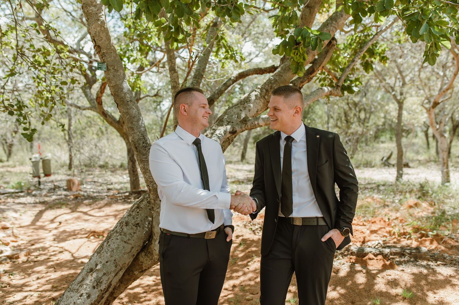 Two men in formal attire shaking hands outdoors under trees at a wedding celebration.