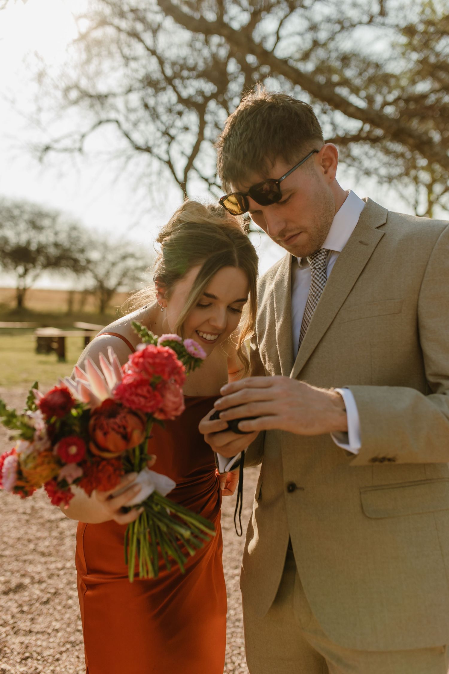 A couple in a tan suit and red dress share an intimate moment during a sunset photoshoot.