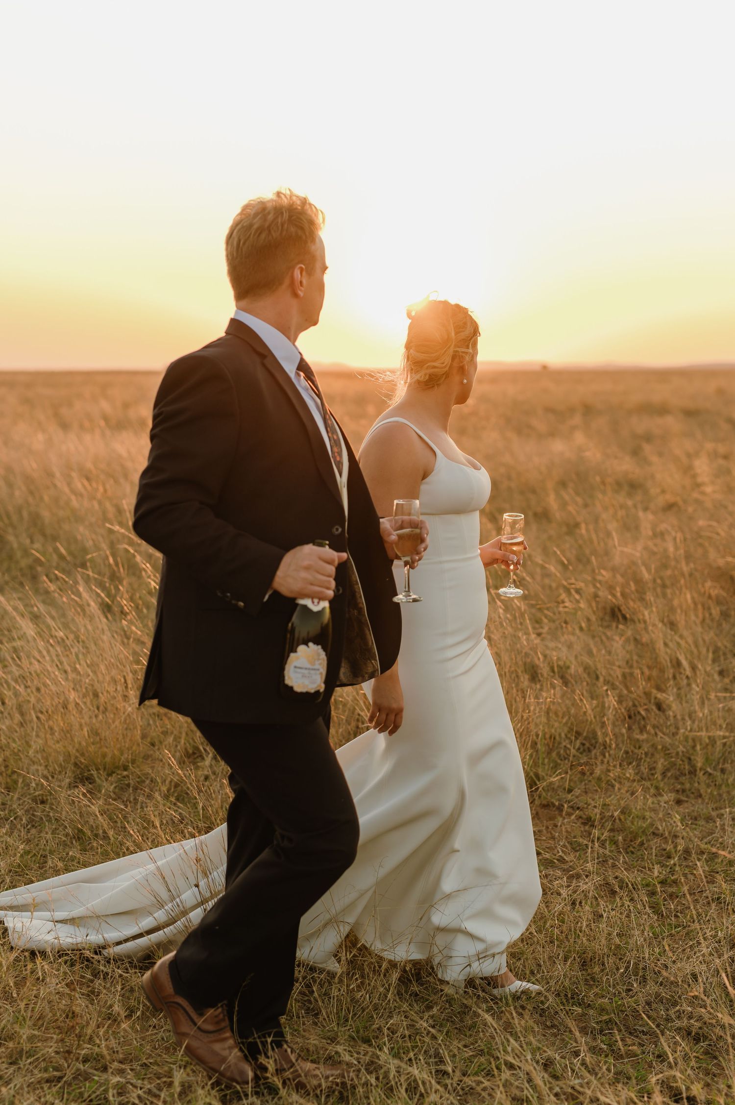 Bride and groom running playfully through tall grass at sunset.