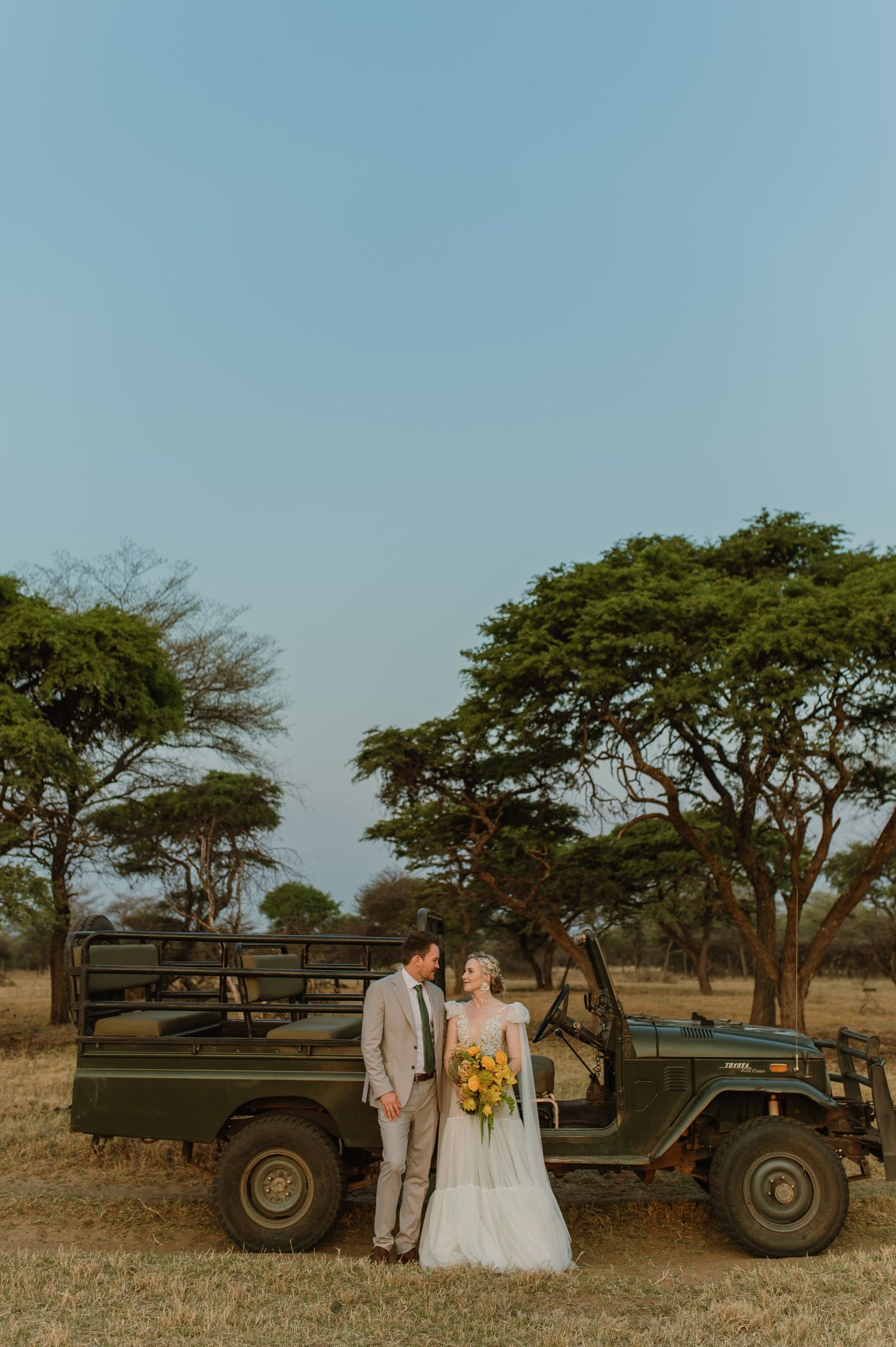 Wedding couple poses with vintage military vehicle under acacia trees in African savanna.