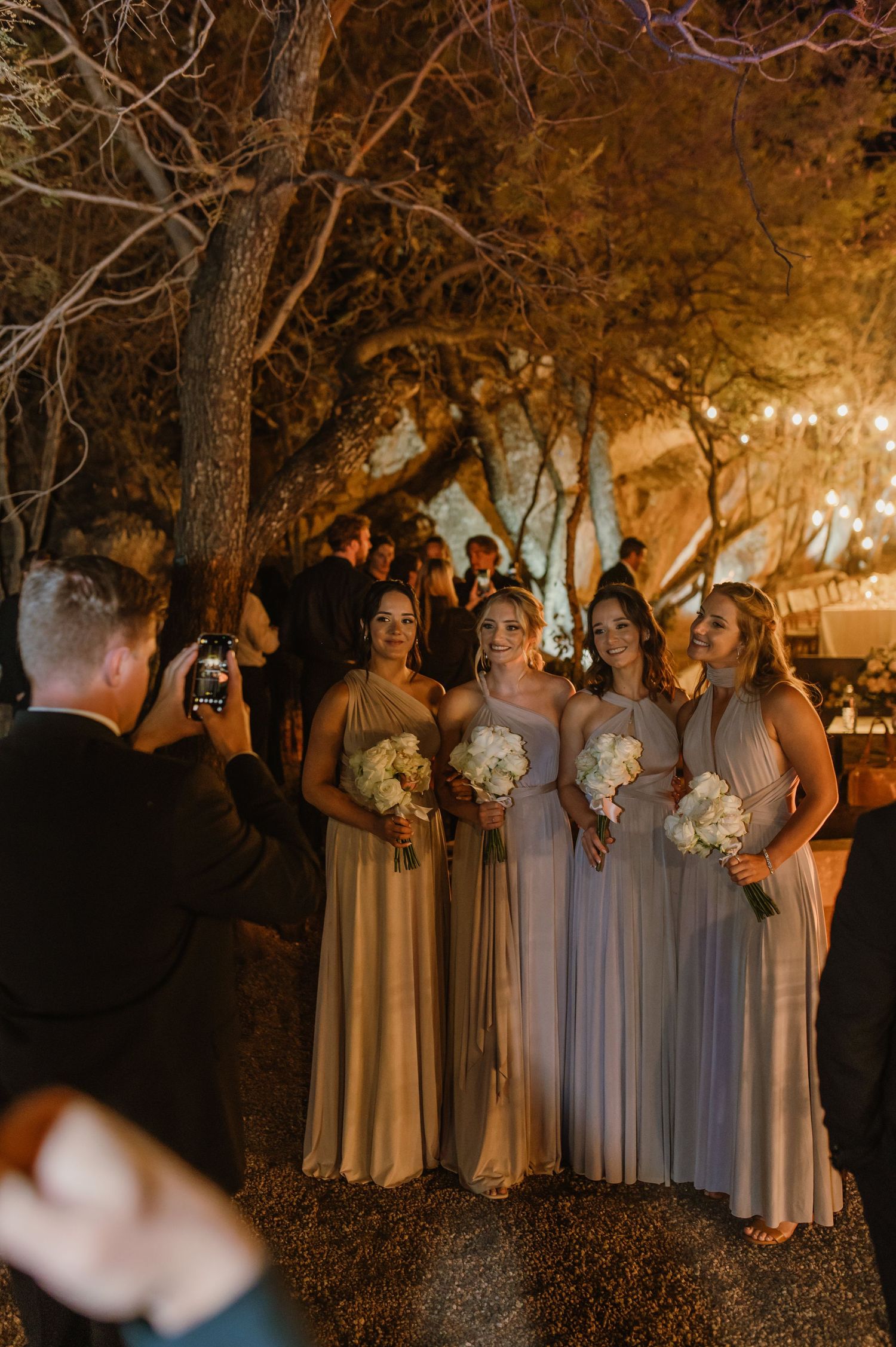 Bridesmaids in light colored dresses pose together under trees at dusk.