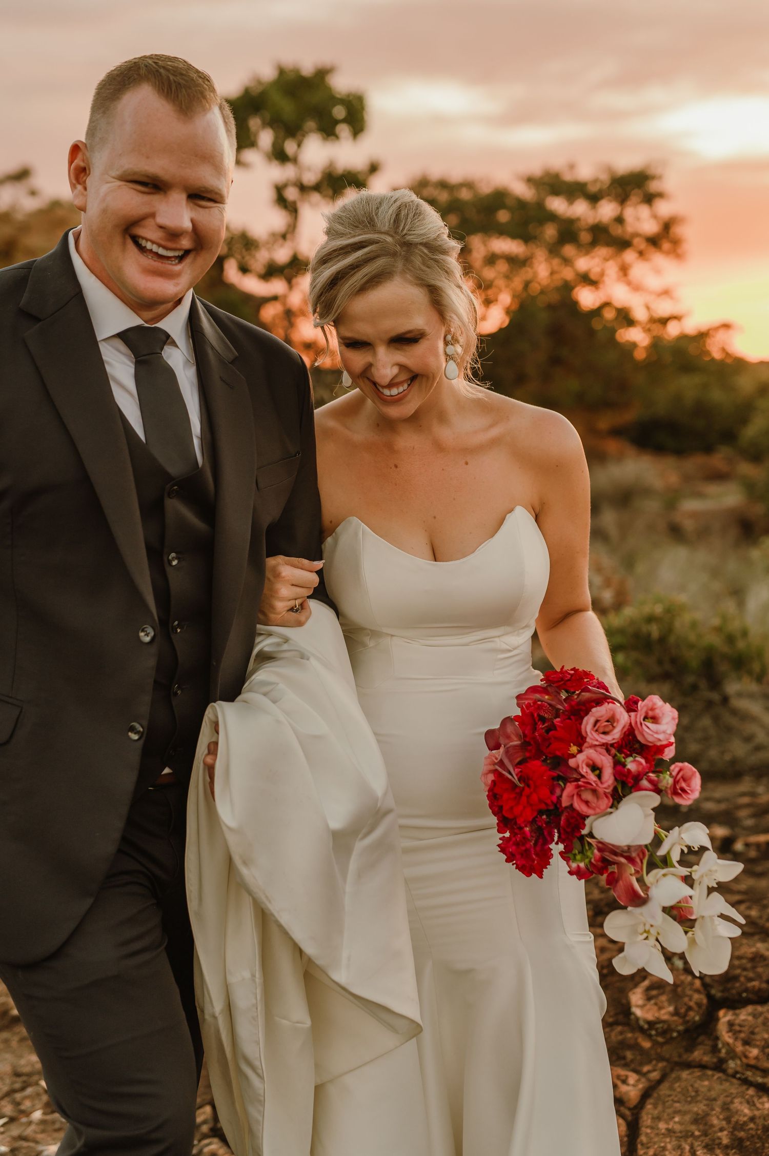Happy wedding couple shares joyful moments with vibrant red bouquet during sunset photoshoot.