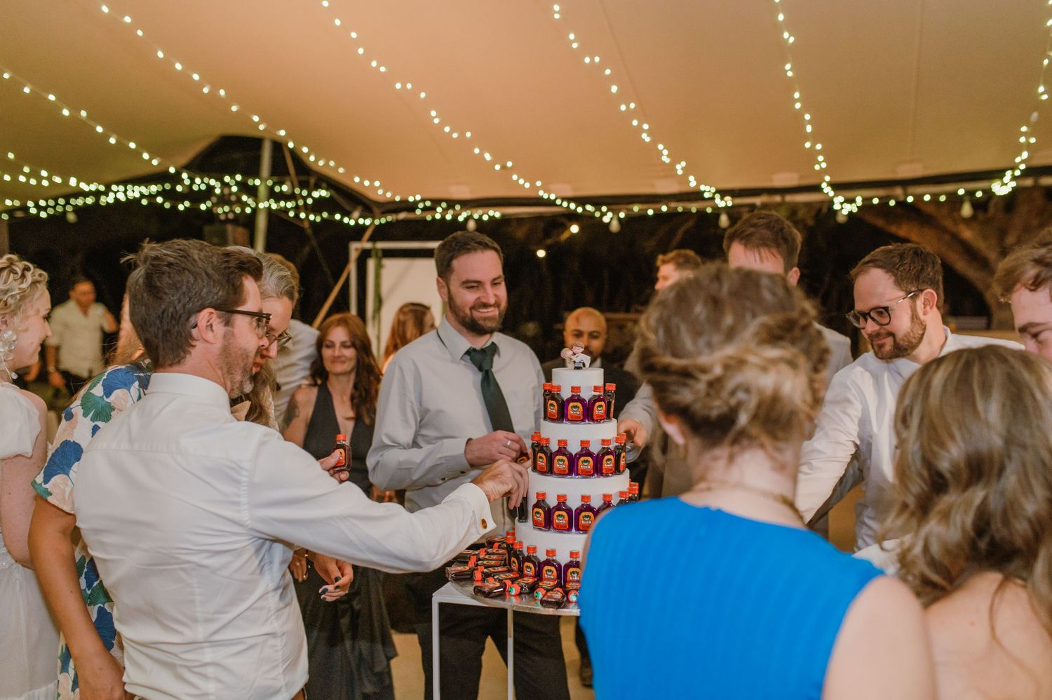 Wedding guests gather around unique Jagermeister bottle cake display at reception.