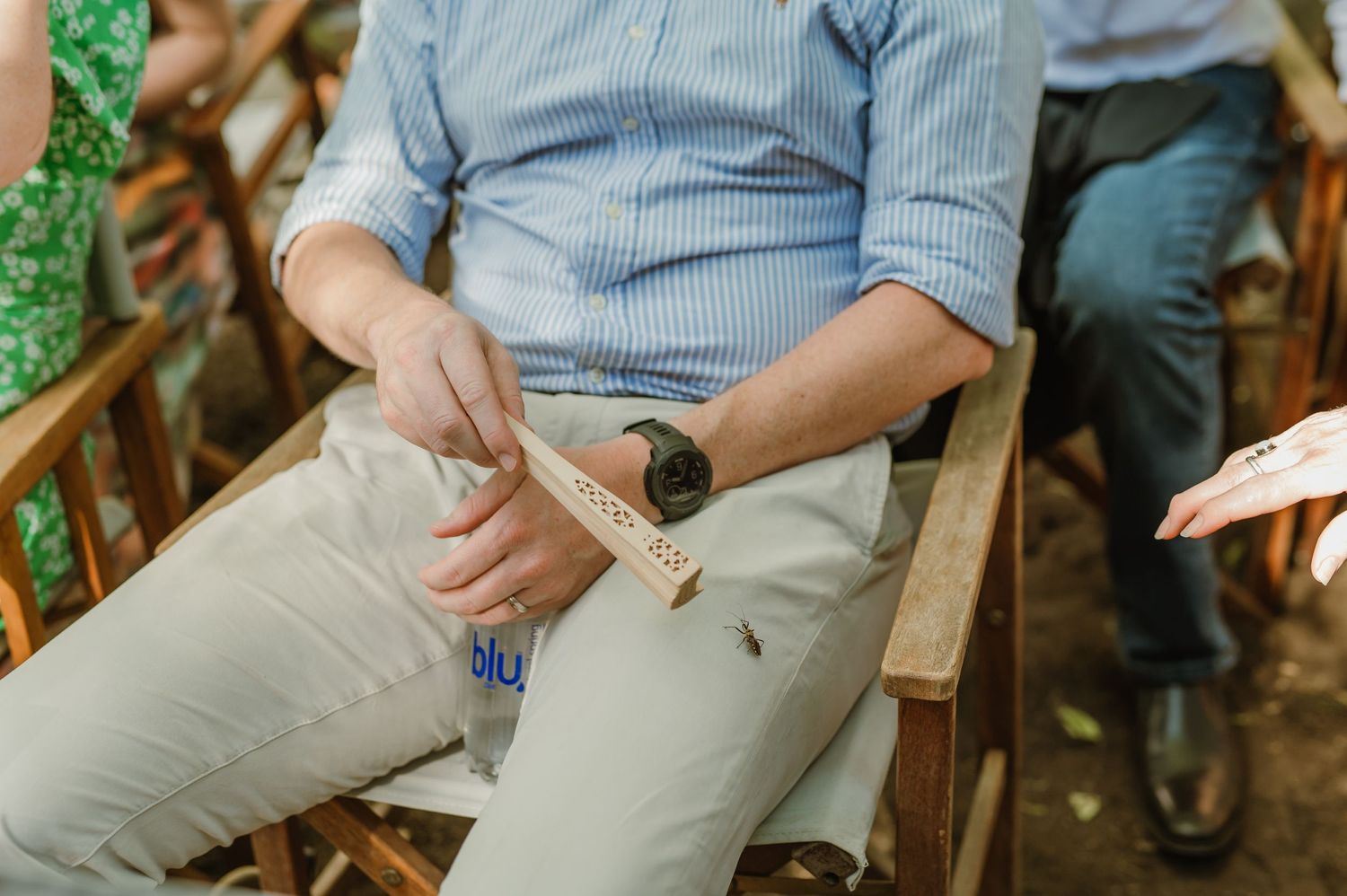 Wedding guests in casual attire seated in wooden chairs during an outdoor ceremony.