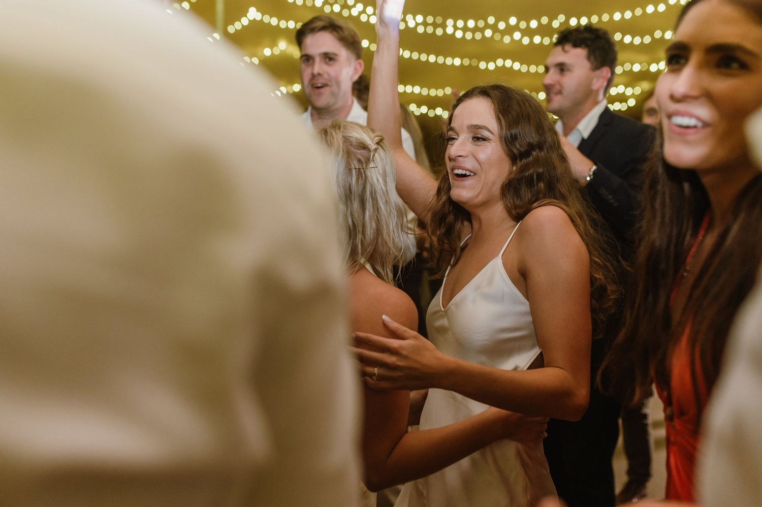 Wedding reception guests laugh and dance together under glowing string lights.